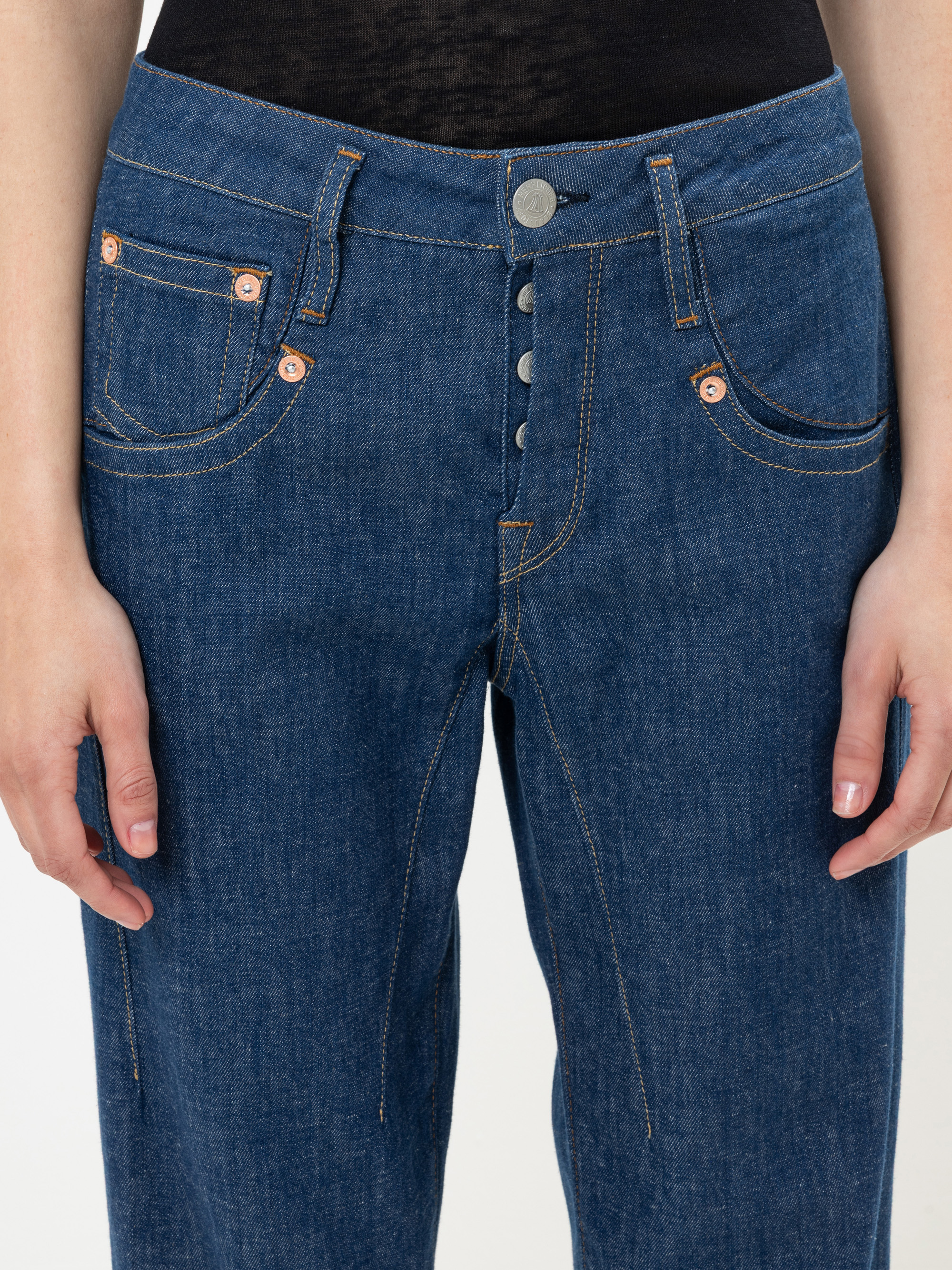 Eine Person trägt eine blaue Denim-Jeans mit sichtbaren Nähten und Metallknöpfen und steht mit den Armen an der Seite. Die Jeans hat einen Hosenschlitz mit Knopf und mehrere Taschen.