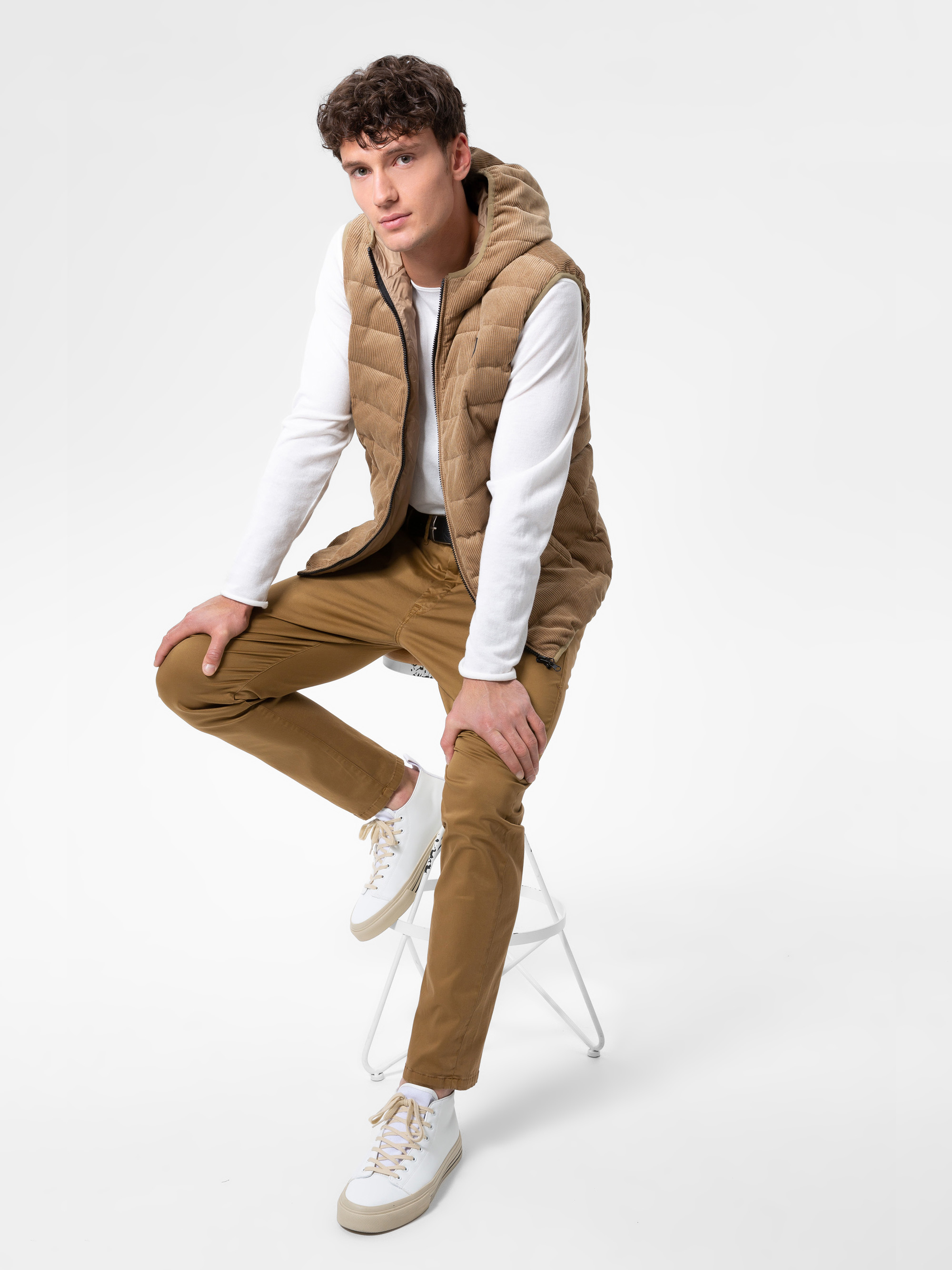 Ein junger Mann mit kurzen lockigen Haaren sitzt auf einem weißen Hocker und trägt eine hellbraune Pufferweste, ein weißes langärmeliges Hemd, eine hellbraune Hose und weiße Turnschuhe vor einem schlichten weißen Hintergrund.