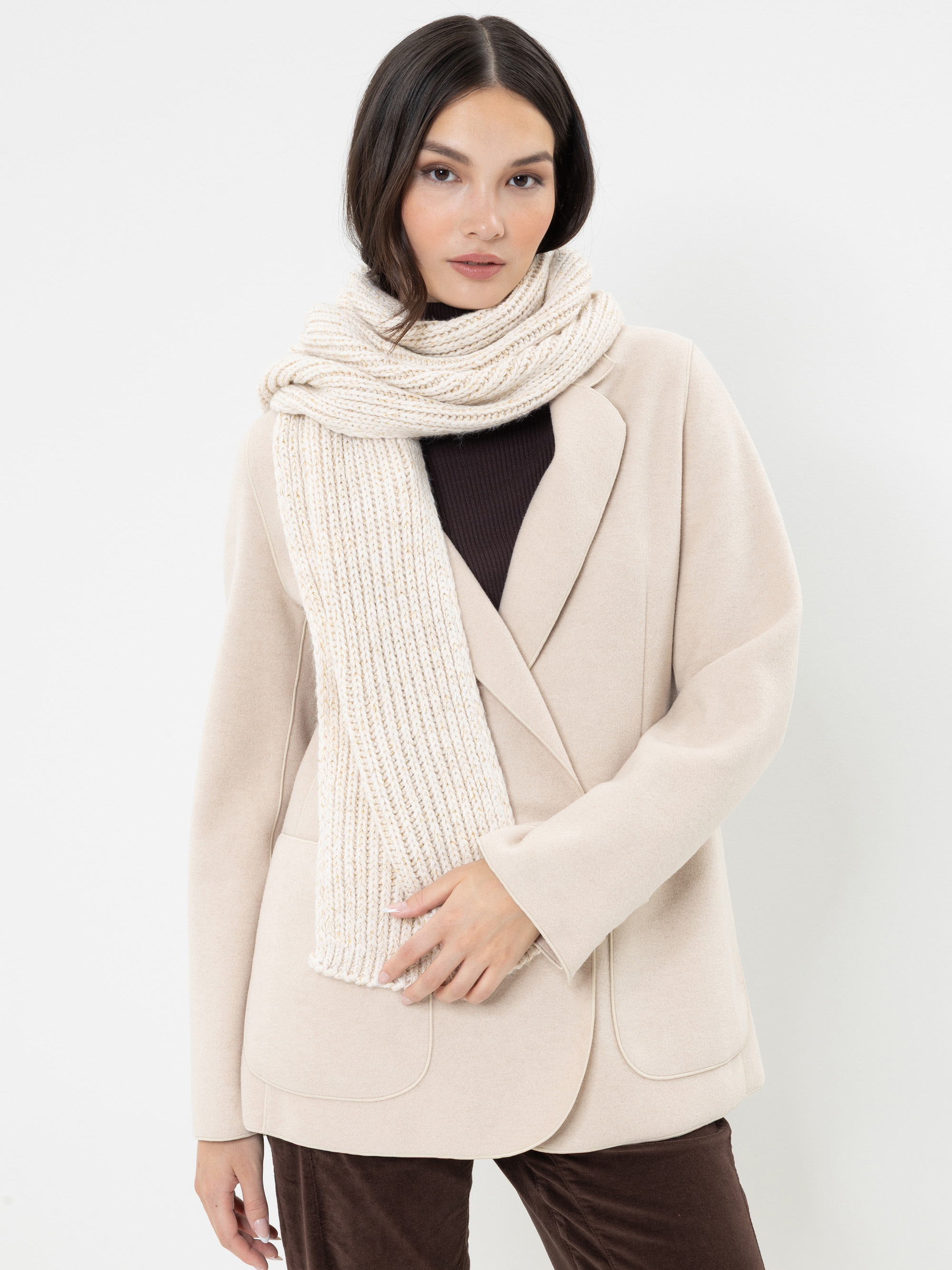 Eine Frau mit langen dunklen Haaren trägt einen cremefarbenen Strickschal, einen beigen Blazer, einen dunklen Rollkragenpullover und eine braune Hose. Sie steht vor einem schlichten weißen Hintergrund und blickt mit ausdruckslosem Gesichtsausdruck in di