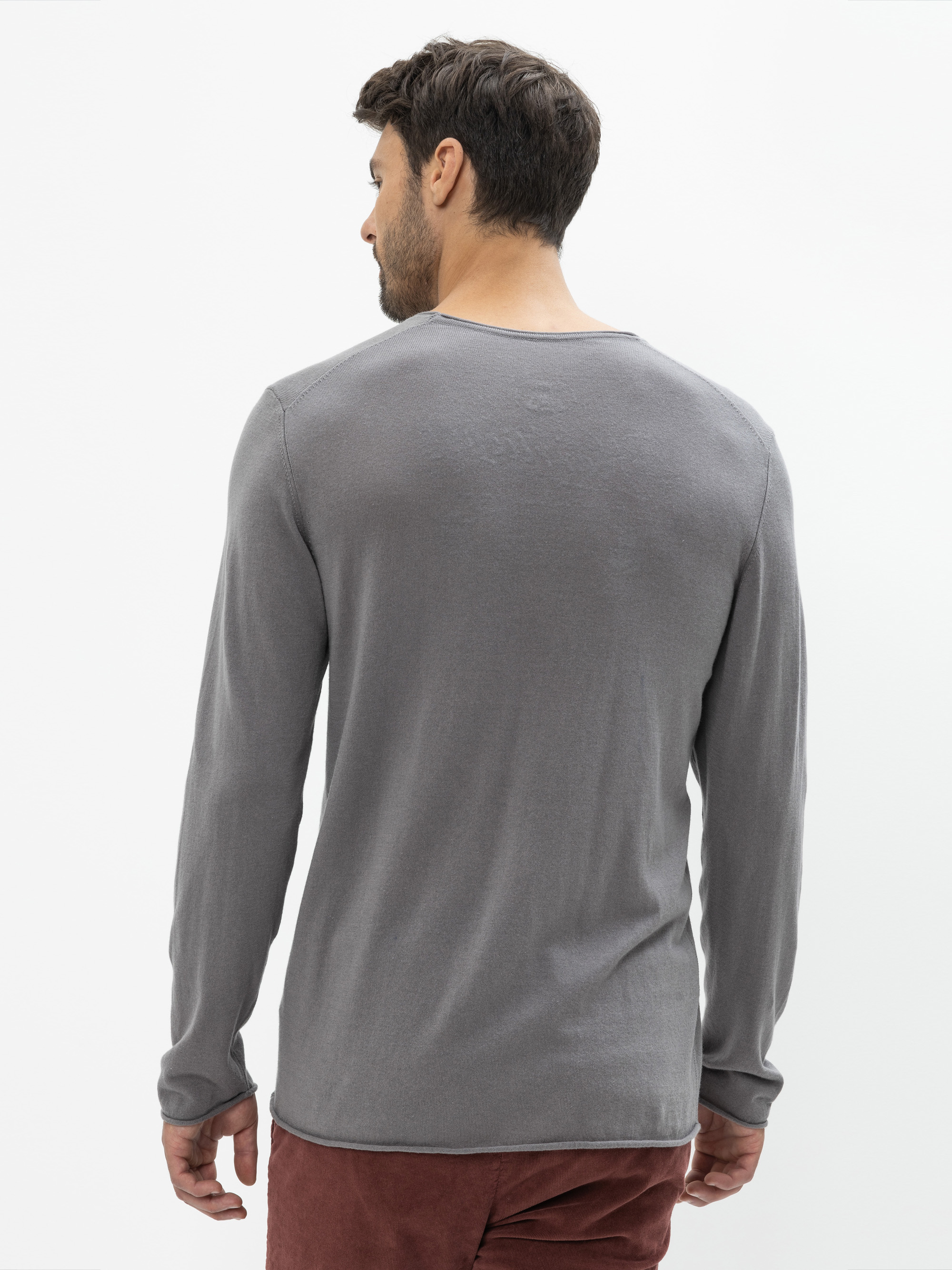 Ein Mann mit kurzen dunklen Haaren wird von hinten gezeigt. Er trägt ein langärmeliges graues Hemd und eine kastanienbraune Hose und steht vor einem schlichten weißen Hintergrund.