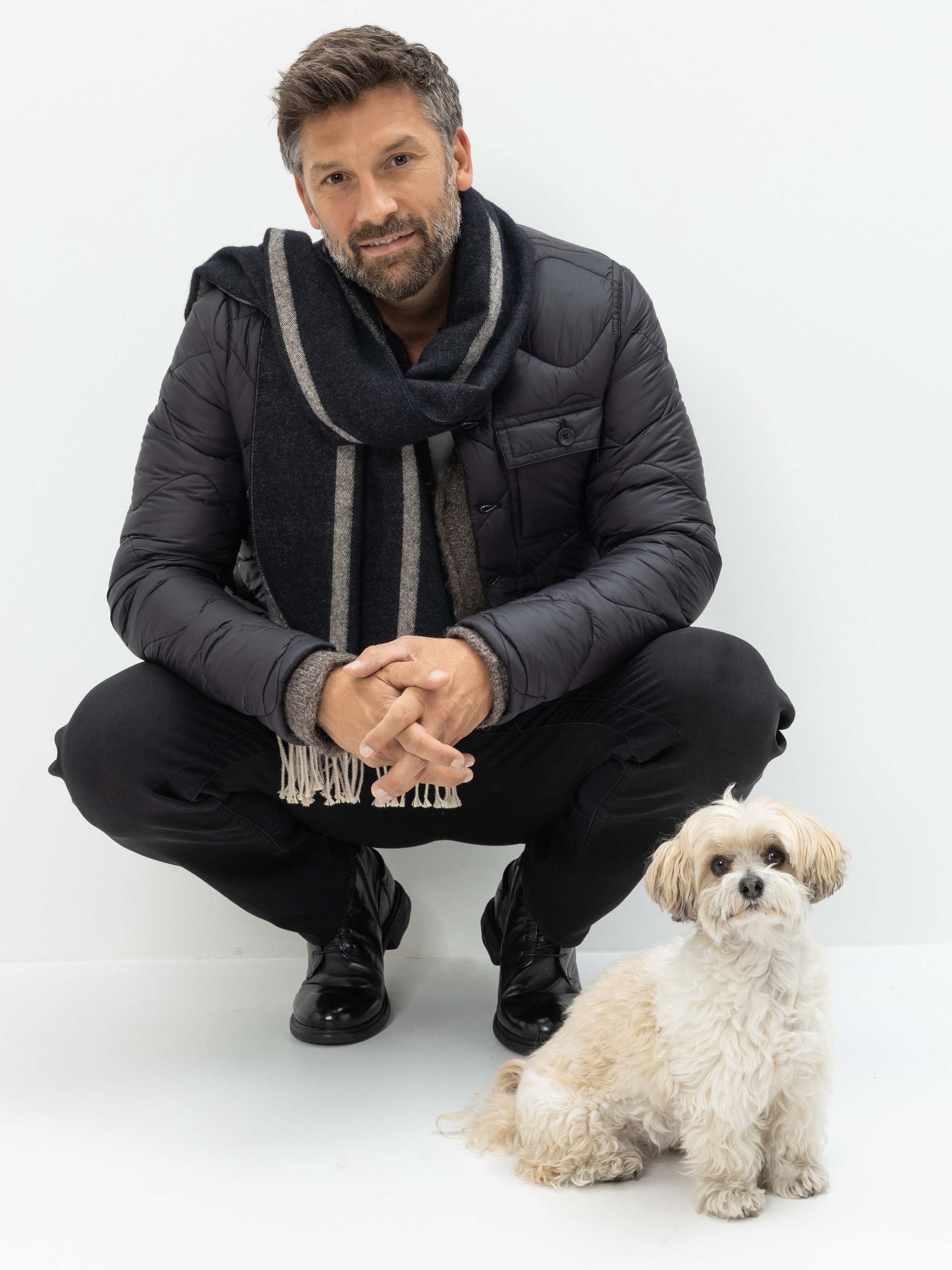 Ein Mann mit dunklem Haar und Bart, der eine schwarze Pufferjacke, einen Schal und eine schwarze Hose trägt, hockt und lächelt in die Kamera. Ein kleiner weiß-hellbrauner Hund sitzt vor ihm auf dem Boden vor einem schlichten weißen Hintergrund.