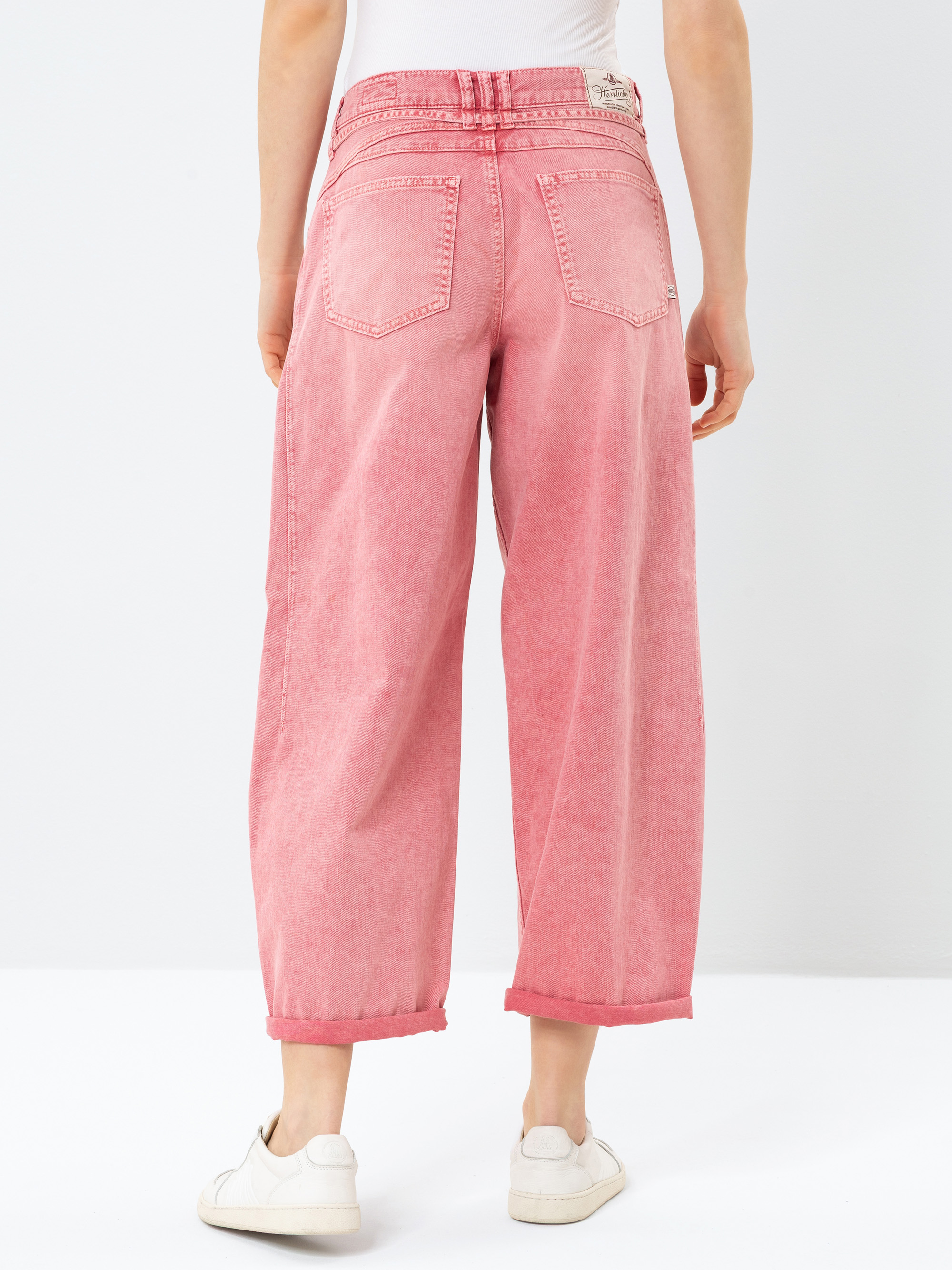 Person, die eine pinkfarbene Jeans mit weitem Bein und aufgerollten Manschetten, ein weißes Hemd und weiße Turnschuhe trägt, von hinten vor einem schlichten weißen Hintergrund.