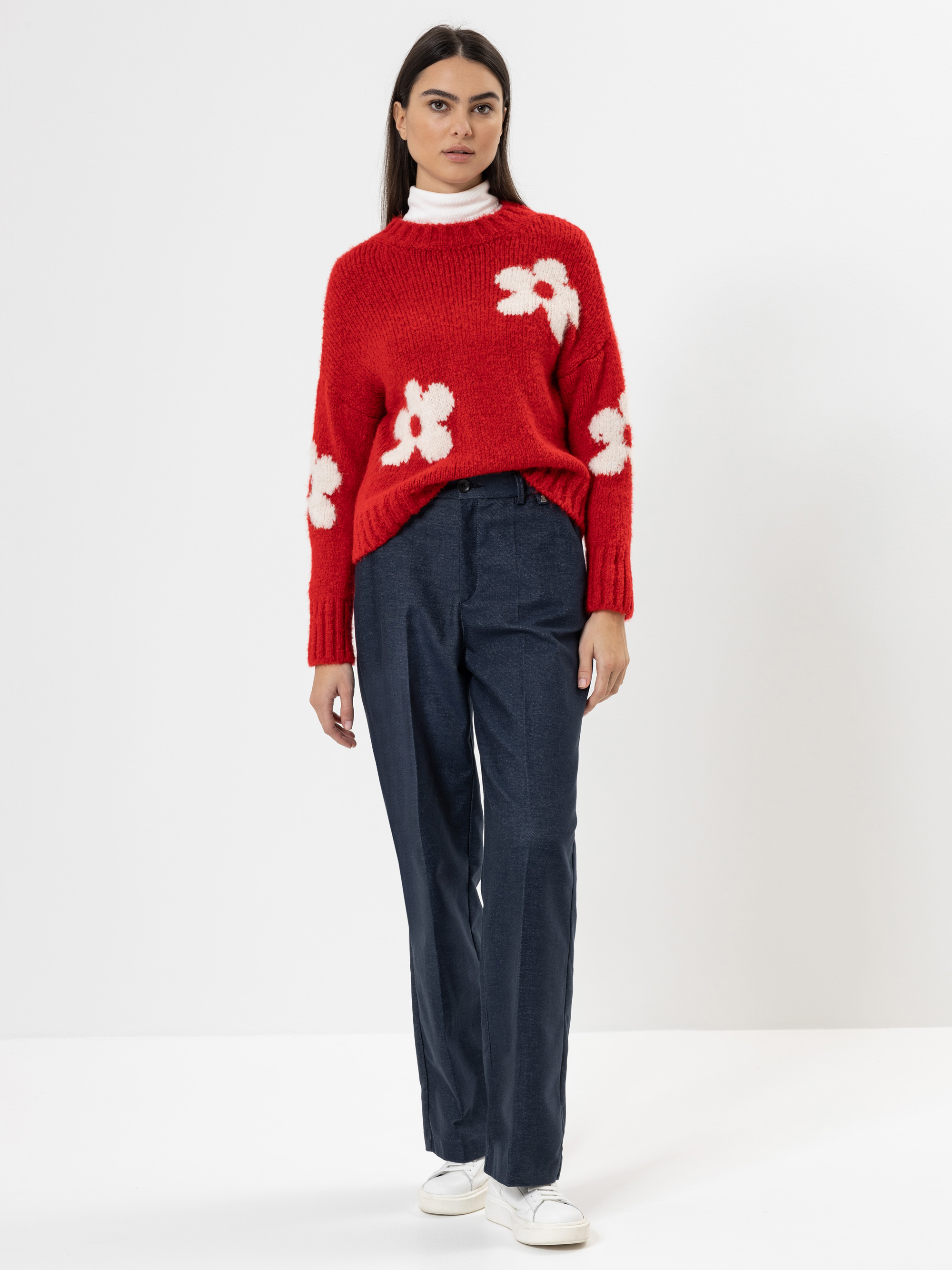 Eine Frau steht vor einem schlichten weißen Hintergrund und trägt einen roten Pullover mit weißen Blumenmustern, eine dunkelblaue Hose und weiße Turnschuhe. Sie hat glattes dunkles Haar und einen neutralen Gesichtsausdruck.