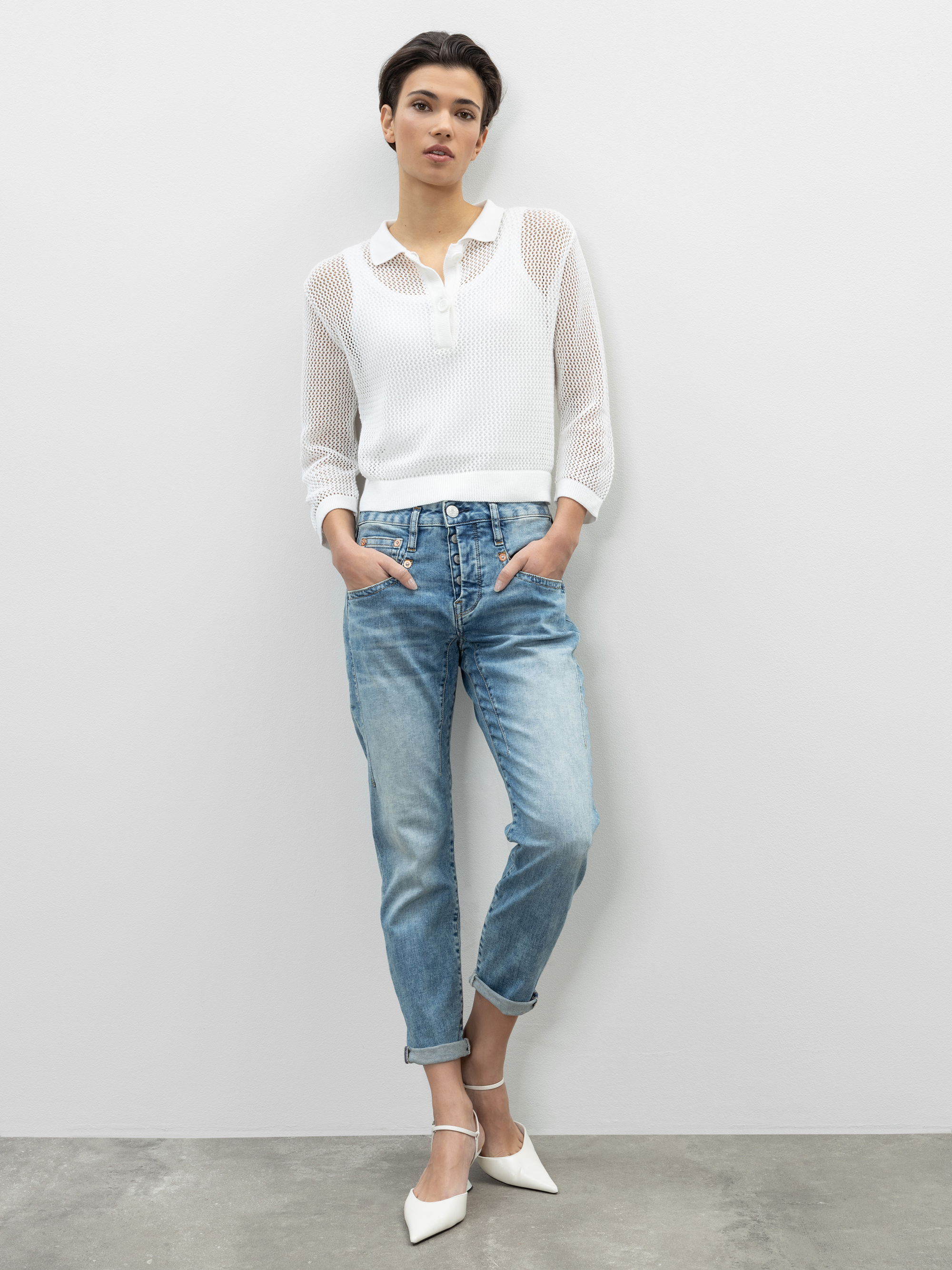 Eine Person steht vor einer schlichten weißen Wand, trägt ein durchsichtiges weißes langärmeliges Oberteil, hellblaue Jeans mit Bündchen und weiße spitze Absätze, die Hände in den Taschen und einen entspannten Gesichtsausdruck.