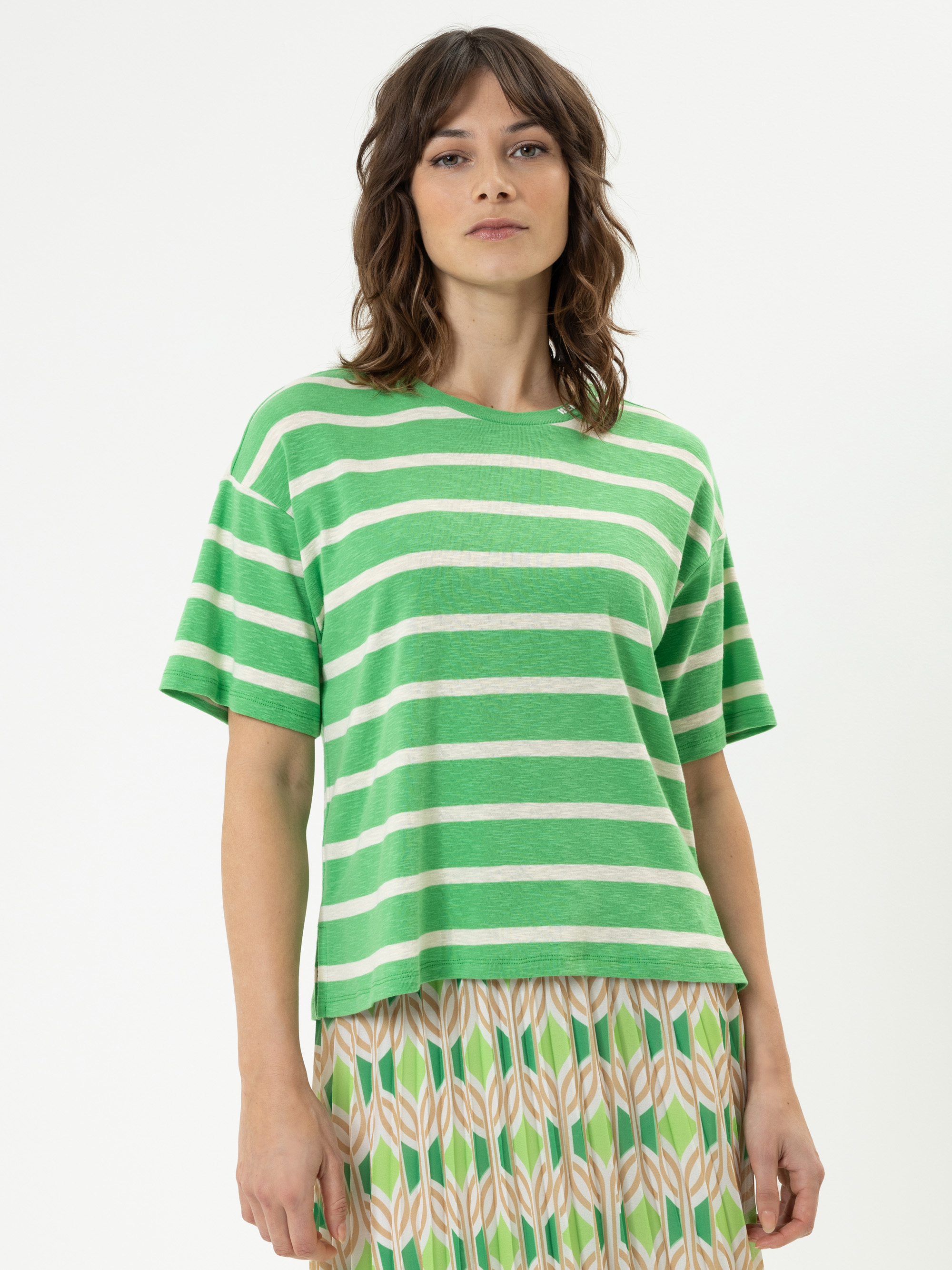 Eine Frau mit gewelltem braunem Haar trägt ein grün-weiß gestreiftes T-Shirt und einen Faltenrock mit einem geometrischen Blattmuster in Beige, Grün und Orange. Sie steht vor einem schlichten weißen Hintergrund.
