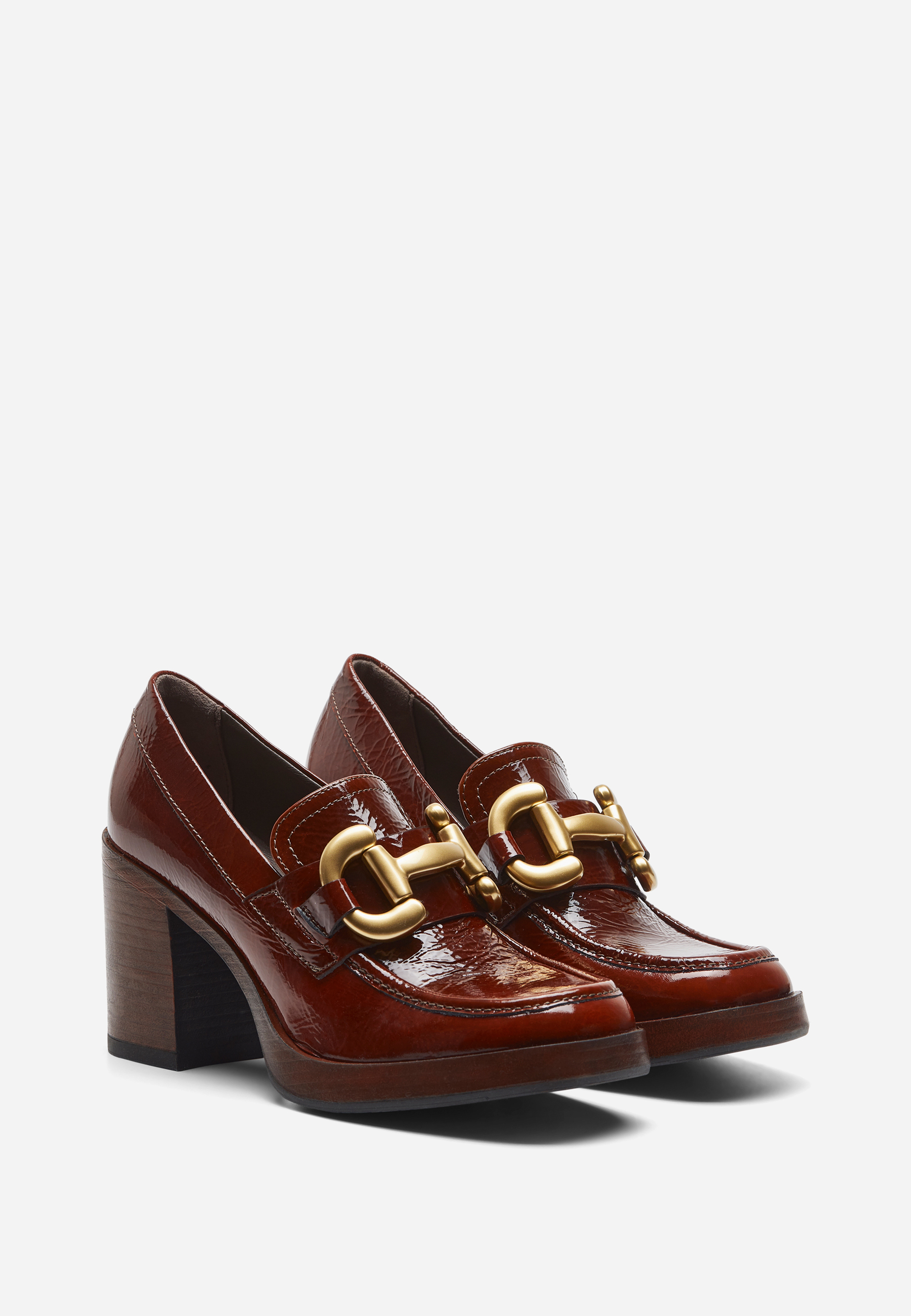 Ein Paar braune Lackleder-Slipper mit Blockabsatz aus Holz und großen goldfarbenen Kettenschnallen auf der Vorderseite, die vor einem schlichten weißen Hintergrund stehen.