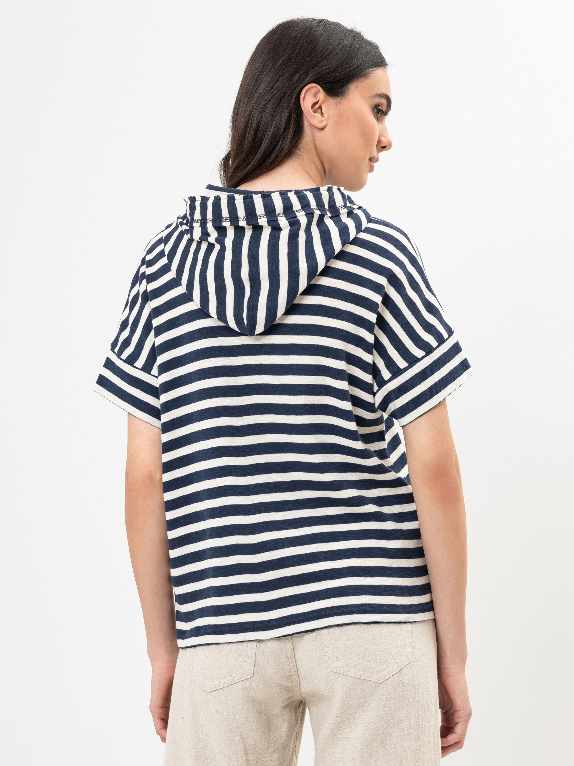 Eine Frau mit dunklem Haar, die einen marineblau-weiß gestreiften kurzärmeligen Kapuzenpullover und eine hellbeige Hose trägt, steht mit dem Rücken zur Kamera vor einem schlichten weißen Hintergrund.