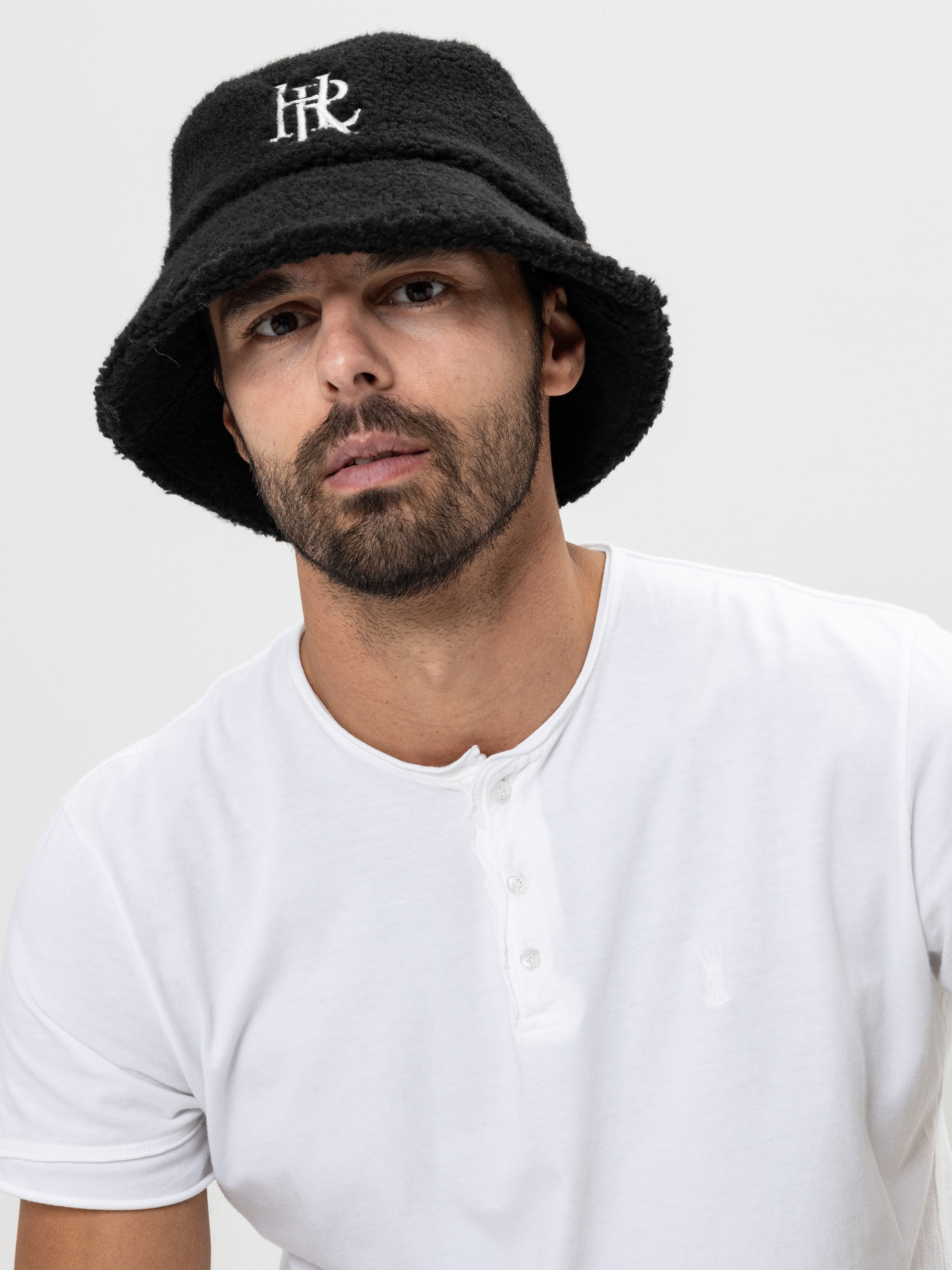 Ein Mann mit kurzem Bart trägt einen schwarzen Eimerhut mit weißer Aufschrift und ein weißes Hemd und posiert vor einem schlichten hellen Hintergrund.