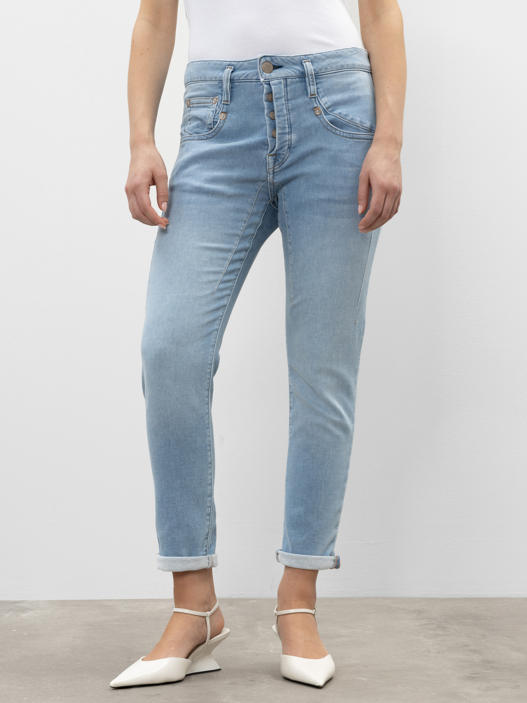 Eine Person in hellblauen, hochgekrempelten Jeans und weißen spitzen Absätzen steht vor einem schlichten weißen Hintergrund. Das Bild ist von den Schultern bis zu den Füßen abgeschnitten.