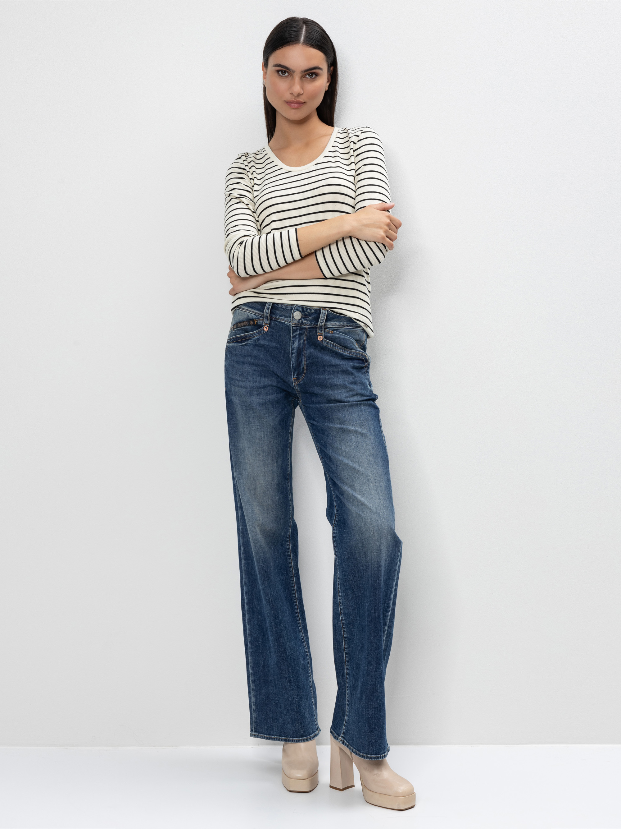 Eine Frau steht mit verschränkten Armen vor einer weißen Wand. Sie trägt ein langärmeliges gestreiftes Hemd, blaue Jeans mit weitem Bein und beigefarbene Plateauabsätze.