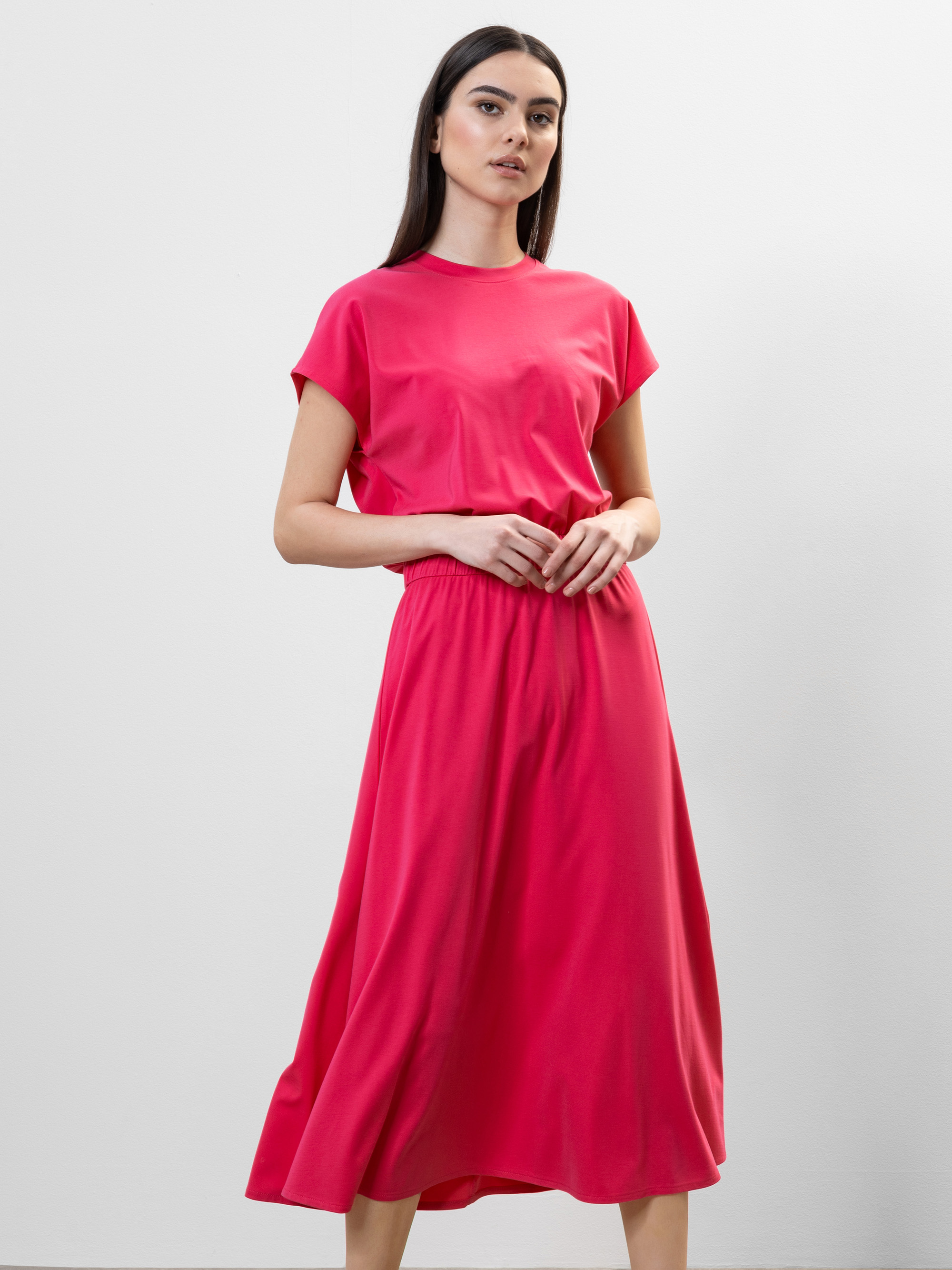 Vor einem schlichten weißen Hintergrund steht eine Frau in einem hellrosa, kurzärmeligen, knöchellangen Kleid mit geraffter Taille und blickt selbstbewusst in die Kamera, während sie die Hände verschränkt.