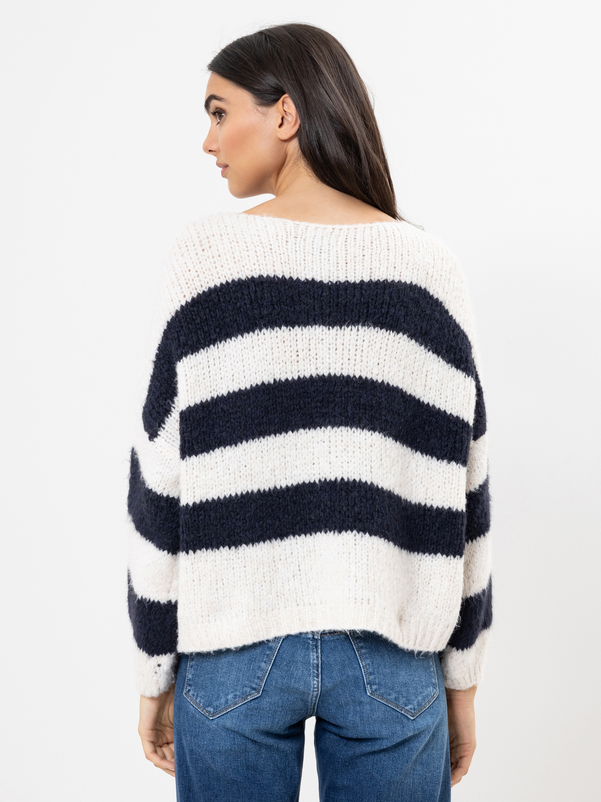 Eine Frau mit langen dunklen Haaren wird von hinten gezeigt. Sie trägt einen weißen Pullover mit dicken schwarzen Querstreifen und blaue Jeans und steht vor einem schlichten weißen Hintergrund.