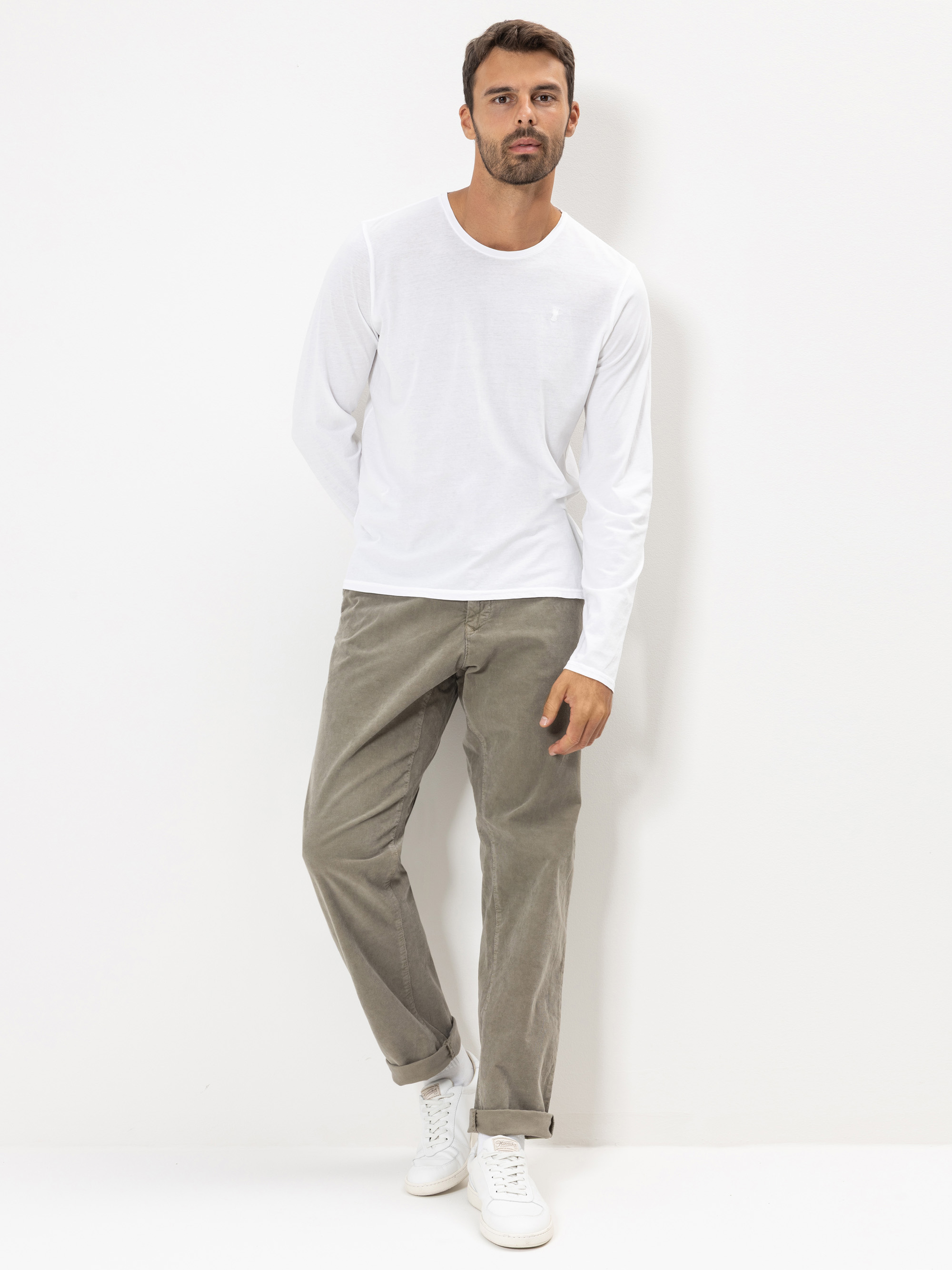 Ein Mann in einem weißen langärmeligen Hemd, einer khakifarbenen Hose und weißen Turnschuhen steht vor einem schlichten weißen Hintergrund, leicht gebeugt, eine Hand in der Tasche, die andere an der Seite.