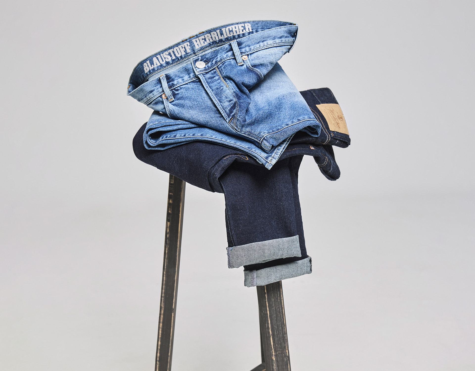 Zwei gefaltete Jeans, eine hellblaue und eine dunkelblaue, sind auf einem Holzschemel gestapelt. Der Bund der oberen Jeans trägt die Aufschrift "BLAUSTOFF HERZBLUTCHER.".