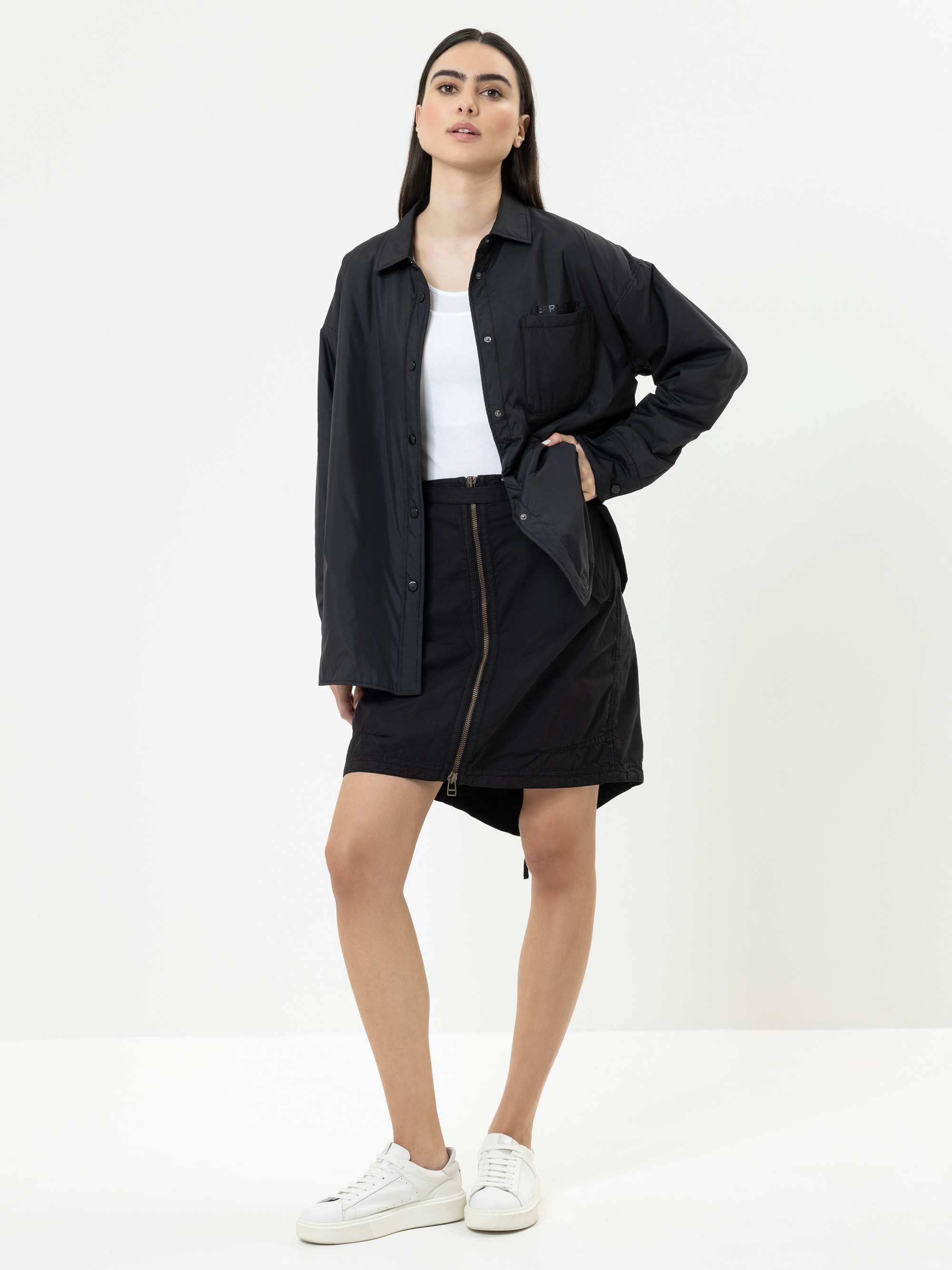 Eine Frau steht vor einem weißen Hintergrund und trägt ein schwarzes Button-up-Hemd, ein weißes Oberteil, einen schwarzen Rock mit einem Reißverschluss und weiße Turnschuhe. Eine Hand ruht auf ihrer Hüfte, während die andere an ihrer Seite hängt.