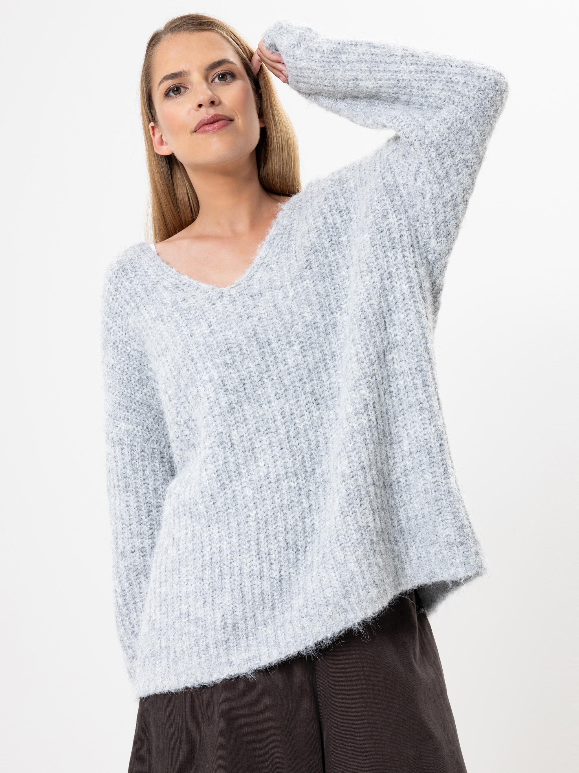 Eine Frau mit langen blonden Haaren trägt einen strukturierten, hellgrauen Pullover mit V-Ausschnitt und eine dunkle Hose. Sie steht vor einem schlichten weißen Hintergrund, hat einen Arm erhoben und einen entspannten, selbstbewussten Gesichtsausdruck.