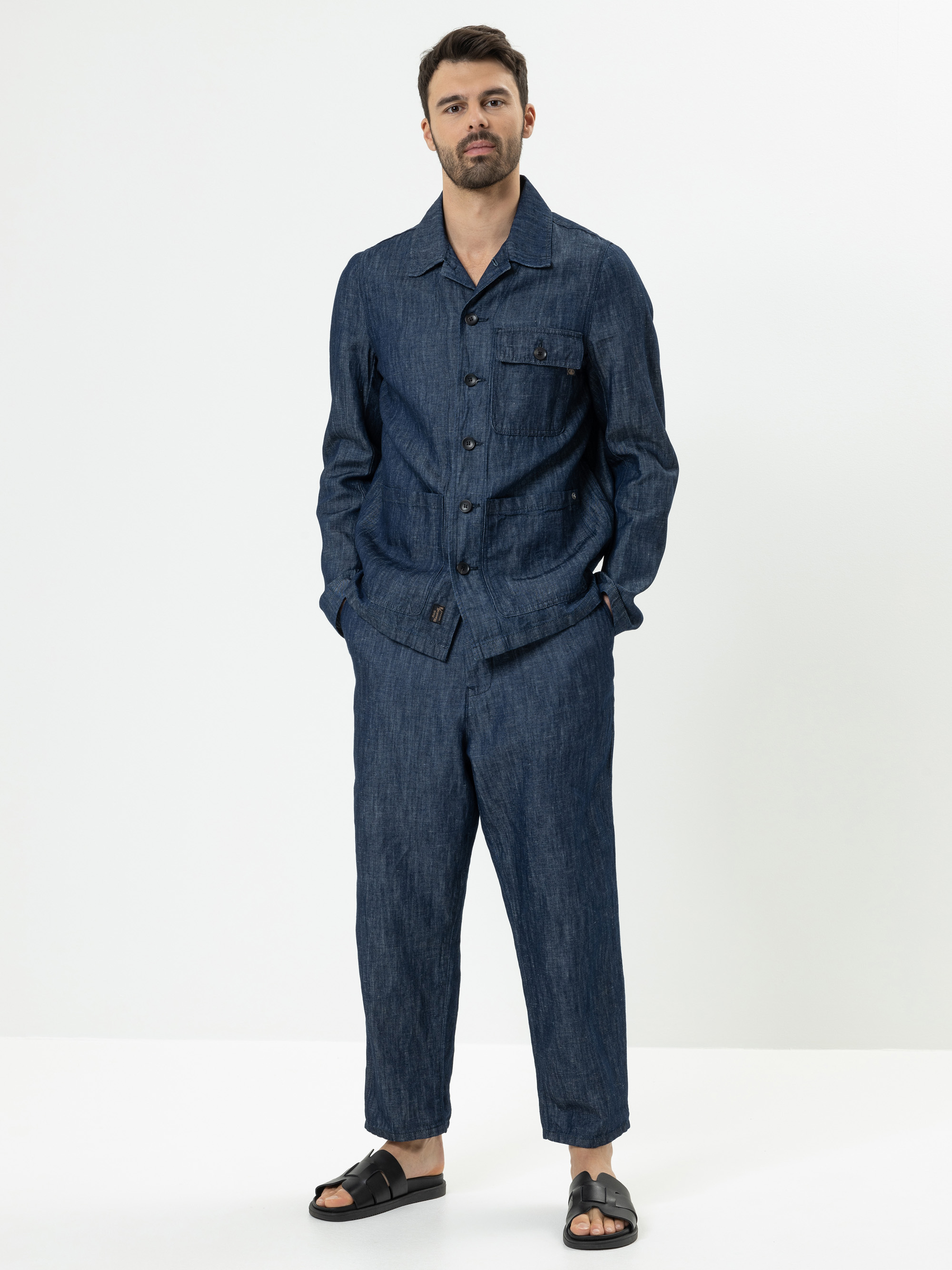 Ein Mann steht vor einem weißen Hintergrund und trägt eine passende dunkelblaue Jeansjacke und eine Hose mit schwarzen Sandalen. Er hat die Hände in den Taschen und trägt kurzes dunkles Haar und einen gestutzten Bart.