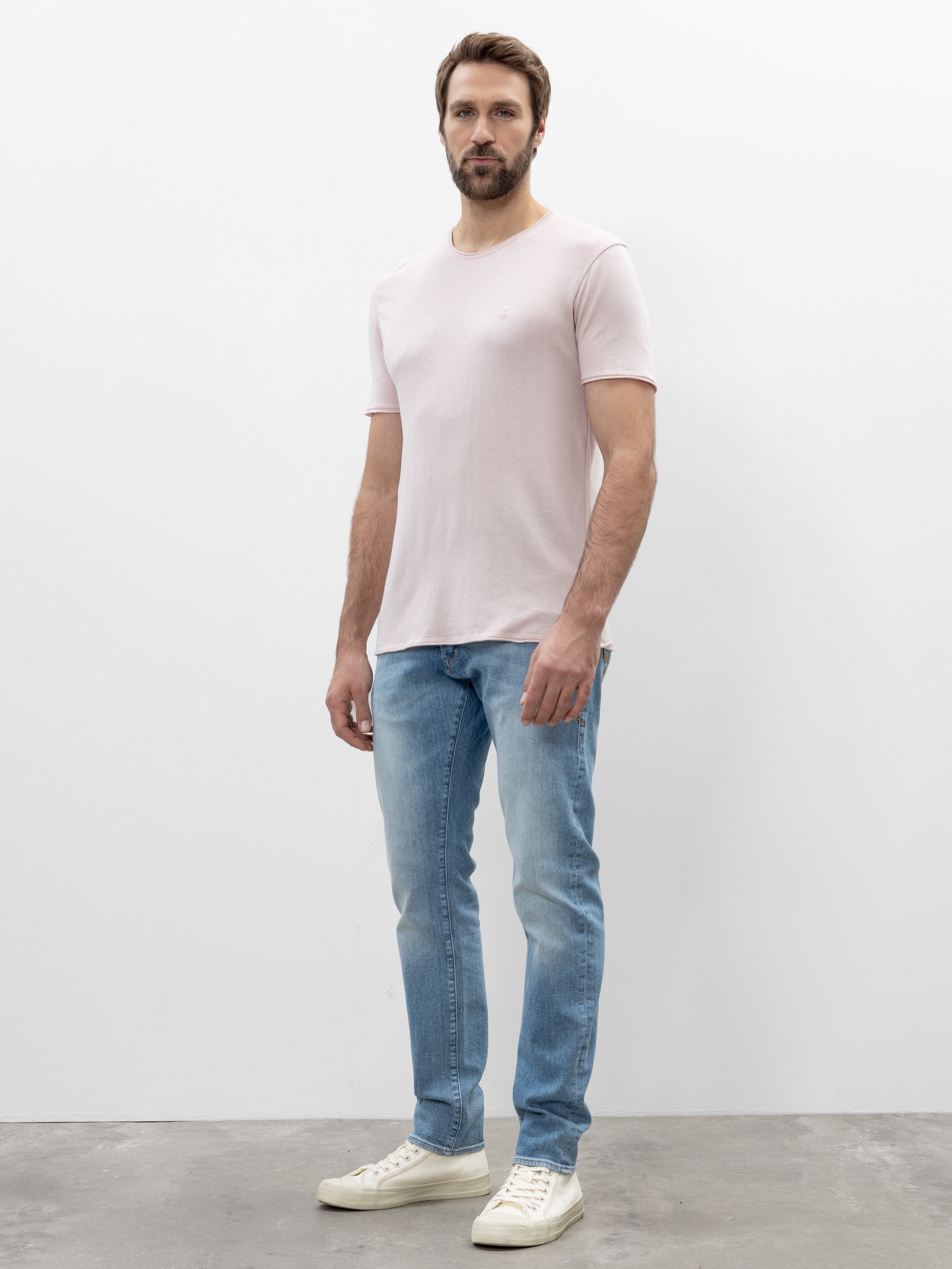 Ein Mann mit kurzen braunen Haaren und einem Bart steht vor einer schlichten weißen Wand und trägt ein hellrosa T-Shirt, hellblaue Jeans und weiße Turnschuhe. Er steht auf einem grauen Boden und blickt leicht nach rechts.