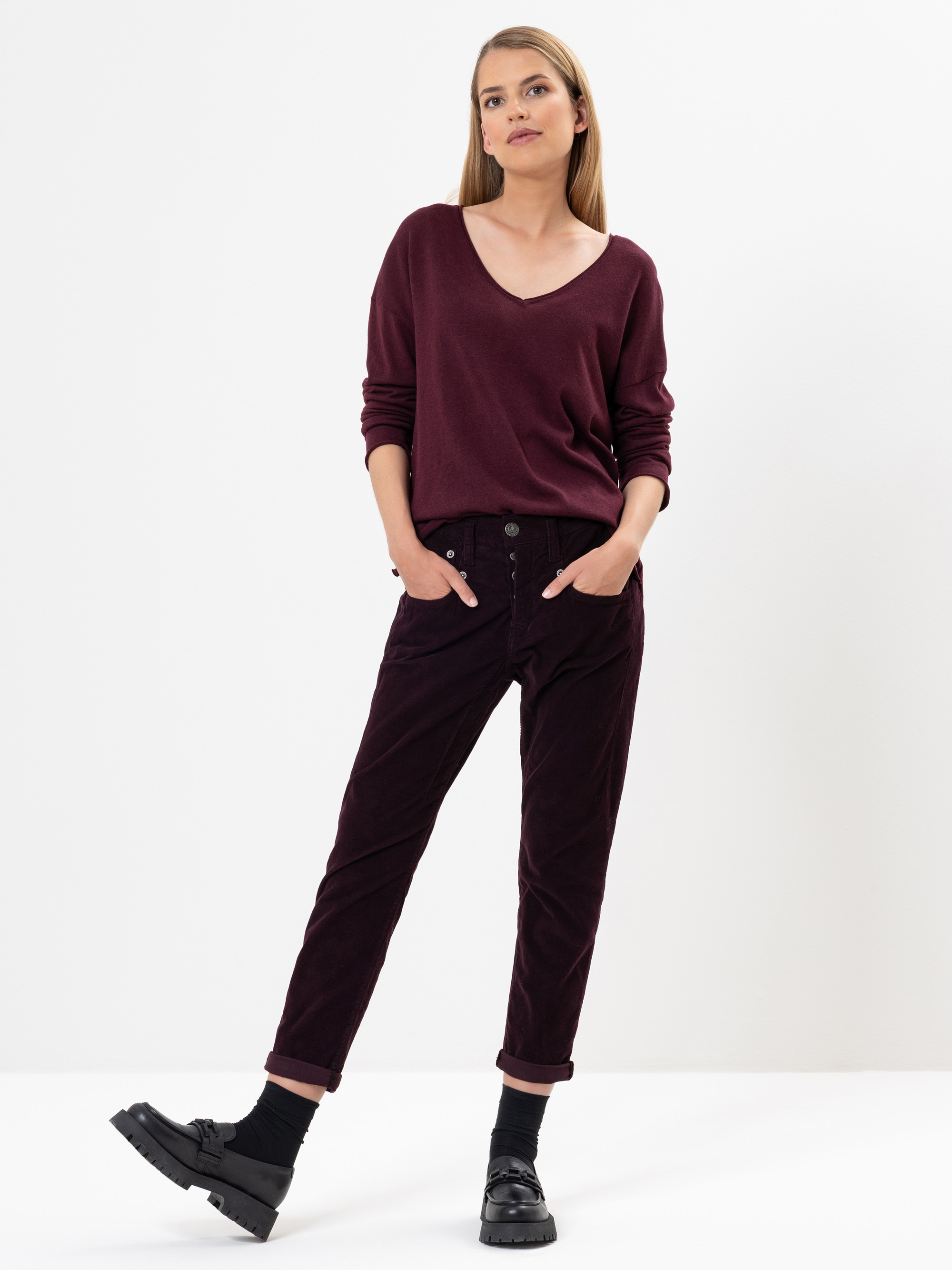 Eine Frau steht vor einem weißen Hintergrund, trägt einen lockeren kastanienbraunen Pullover mit V-Ausschnitt, eine hochgekrempelte dunkle Cordhose, schwarze Socken und schwarze Halbschuhe, die Hände in den Taschen und einen entspannten Gesichtsausdruc