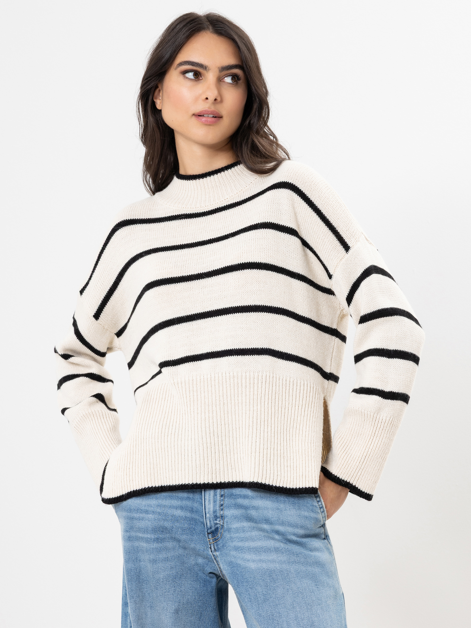 Eine Frau mit langen dunklen Haaren trägt einen cremefarbenen Pullover mit schwarzen Querstreifen und hellblaue Jeans und steht mit den Händen in den Taschen vor einem schlichten weißen Hintergrund.