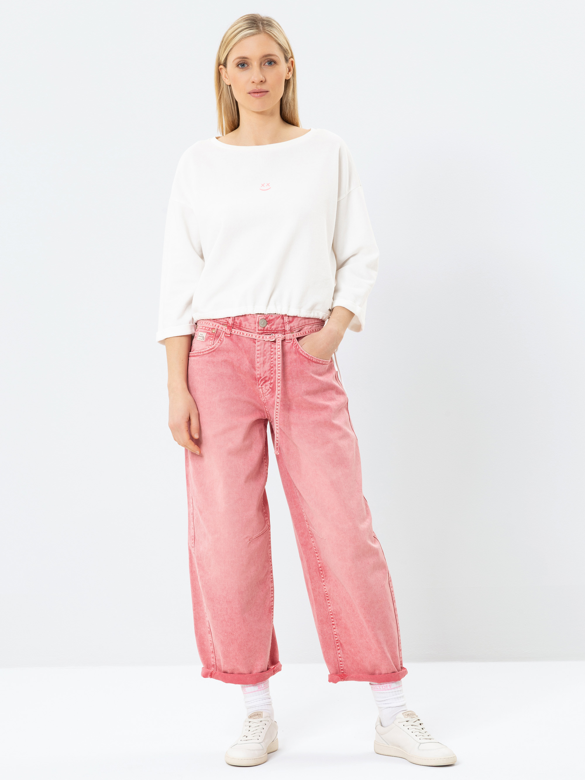 Eine Frau steht vor einem einfarbig weißen Hintergrund und trägt ein lockeres weißes langärmeliges Oberteil, eine hochgeschnittene, weit geschnittene rosa Jeans, weiße Socken und weiße Turnschuhe. Sie hat eine Hand in der Tasche und einen entspannte