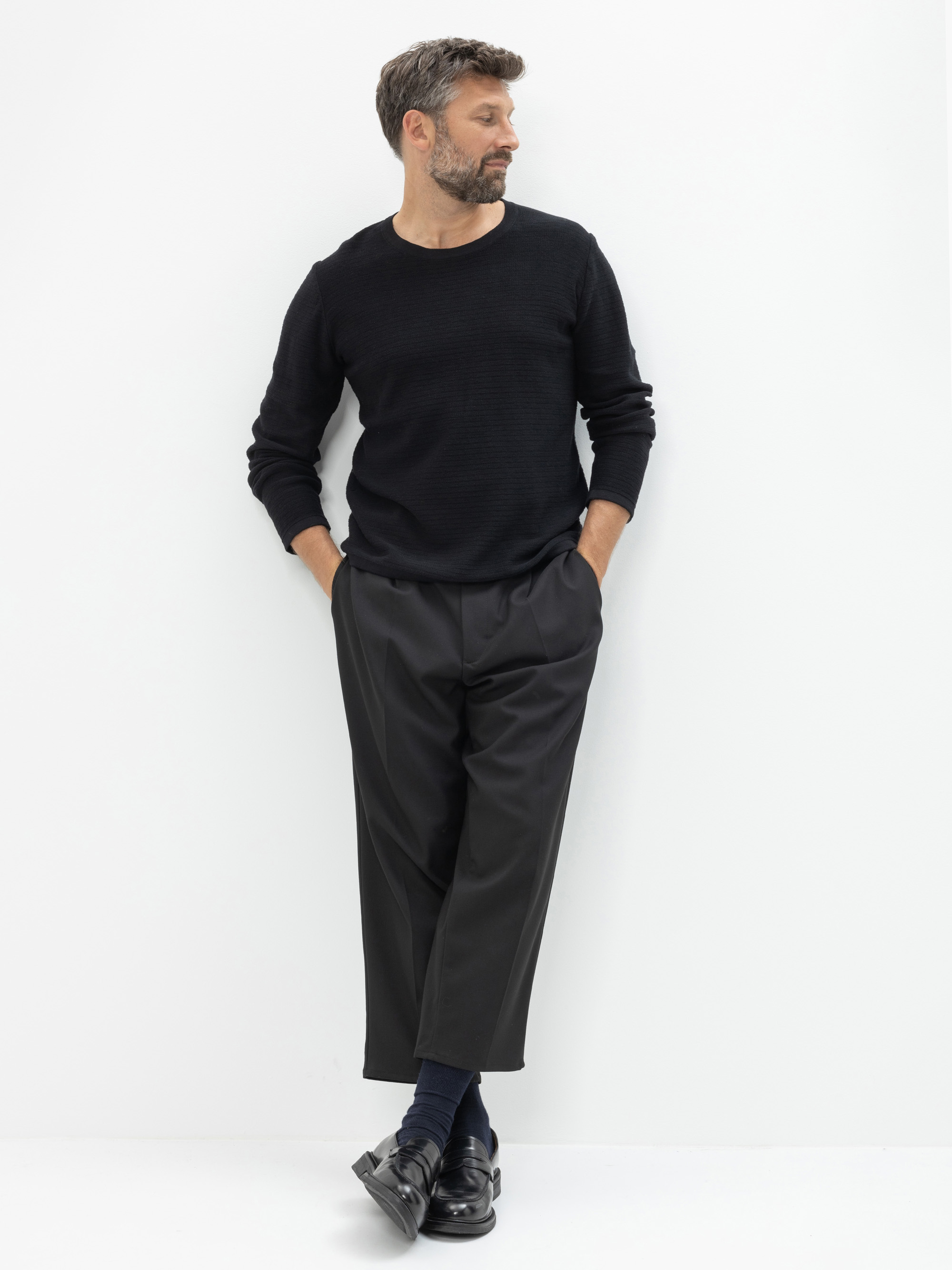 Ein Mann mit kurzen Haaren und Bart steht vor einem weißen Hintergrund. Er trägt ein schwarzes langärmeliges Hemd, eine schwarze kurze Hose, schwarze Socken und schwarze Halbschuhe, die Hände in den Taschen und die Beine übereinander geschlagen.