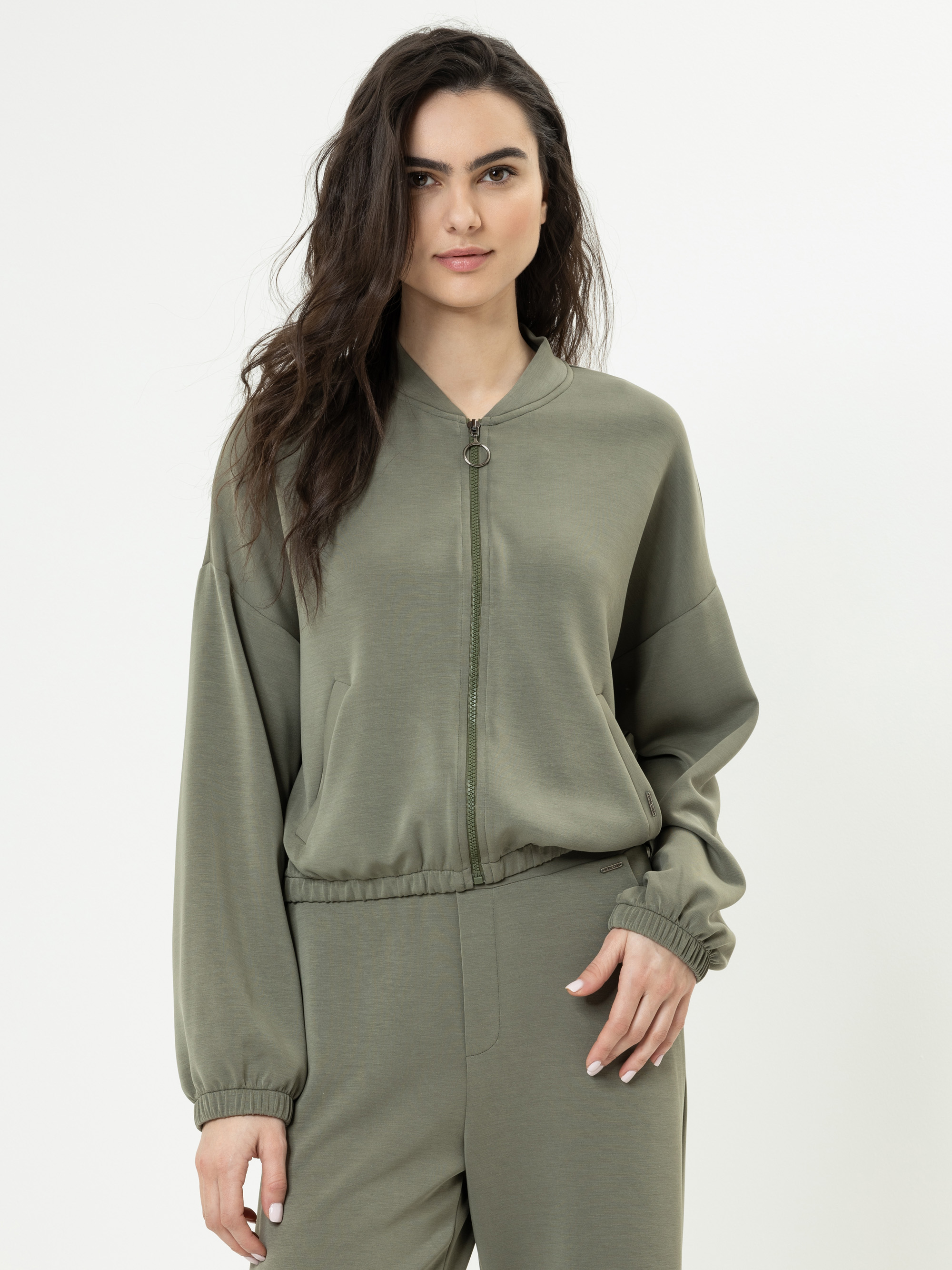 Eine Frau mit langen, gewellten dunklen Haaren trägt eine olivgrüne Jacke mit Reißverschluss und eine passende Hose. Sie steht mit neutralem Gesichtsausdruck vor einem schlichten weißen Hintergrund.