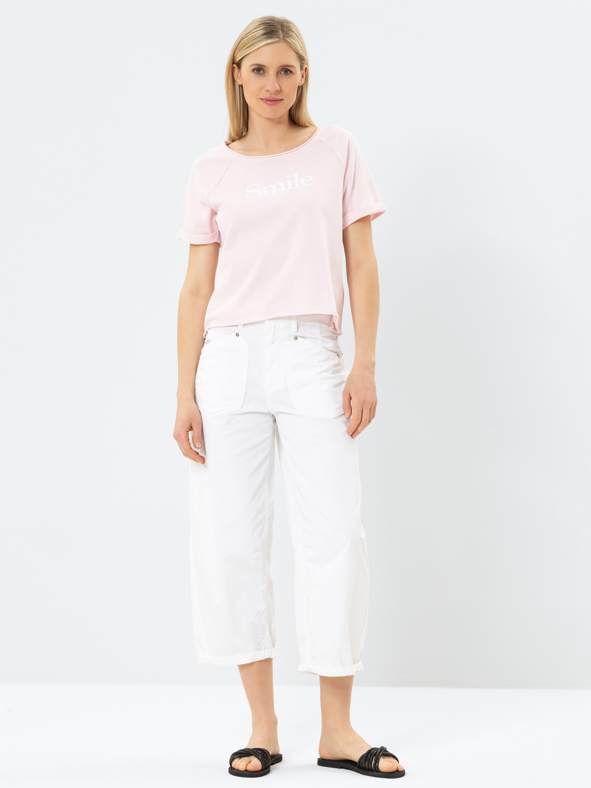 Eine Frau steht vor einem schlichten weißen Hintergrund und trägt ein hellrosa T-Shirt mit der Aufschrift Smile, eine weiße Hose mit weitem Bein und schwarze Slide-Sandalen. Sie hat glattes, blondes Haar und einen entspannten Gesichtsausdruck.
