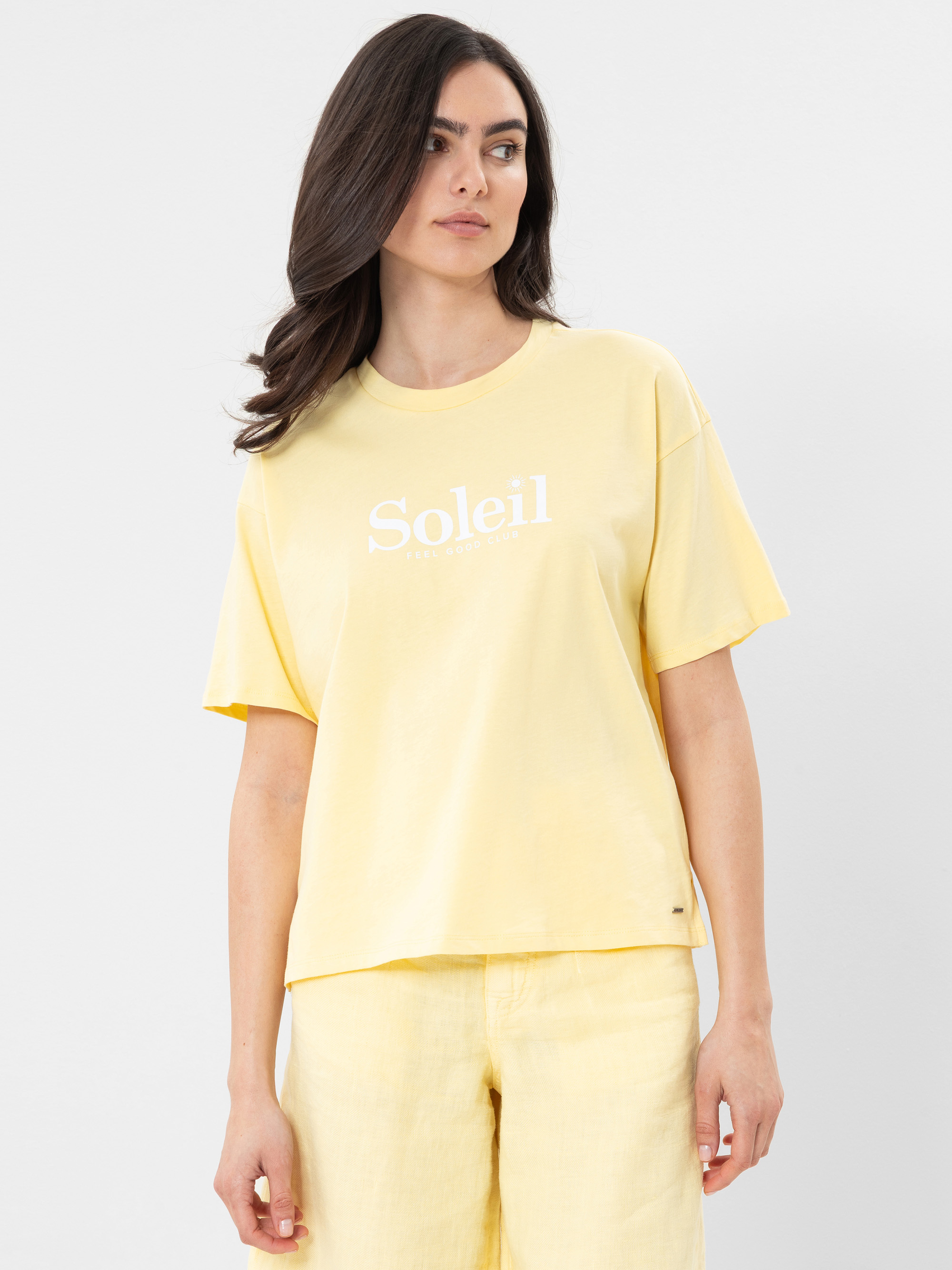 Eine Frau mit langen braunen Haaren trägt ein hellgelbes T-Shirt mit dem Aufdruck "Soleil" und eine dazu passende gelbe Hose und steht vor einem schlichten hellgrauen Hintergrund.