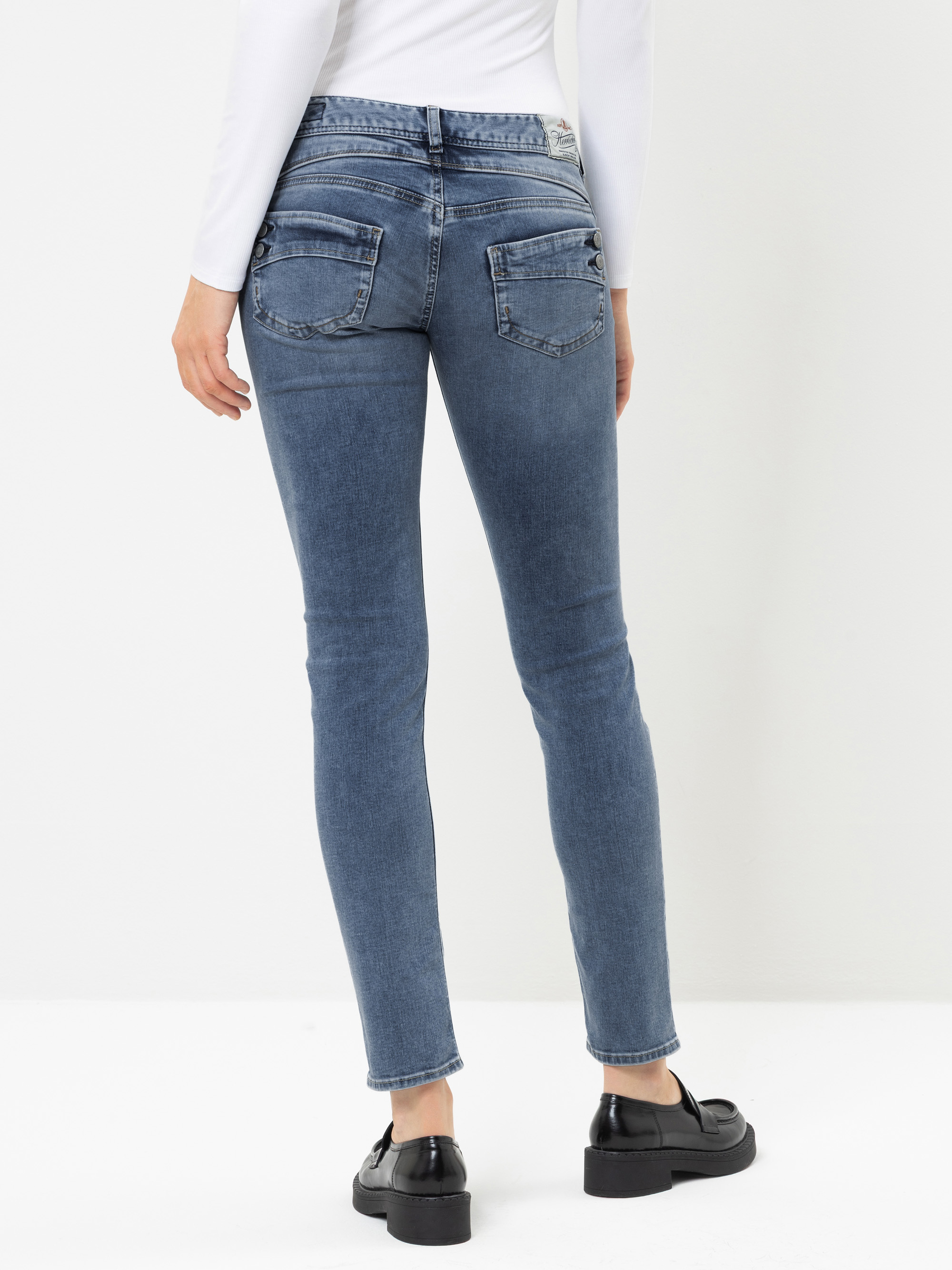 Eine Person, die mit dem Rücken zur Kamera steht und ein weißes Langarmshirt, eine blaue Skinny-Jeans und schwarze Slipper trägt. Der Hintergrund ist schlicht und weiß, wodurch das Outfit besonders gut zur Geltung kommt.