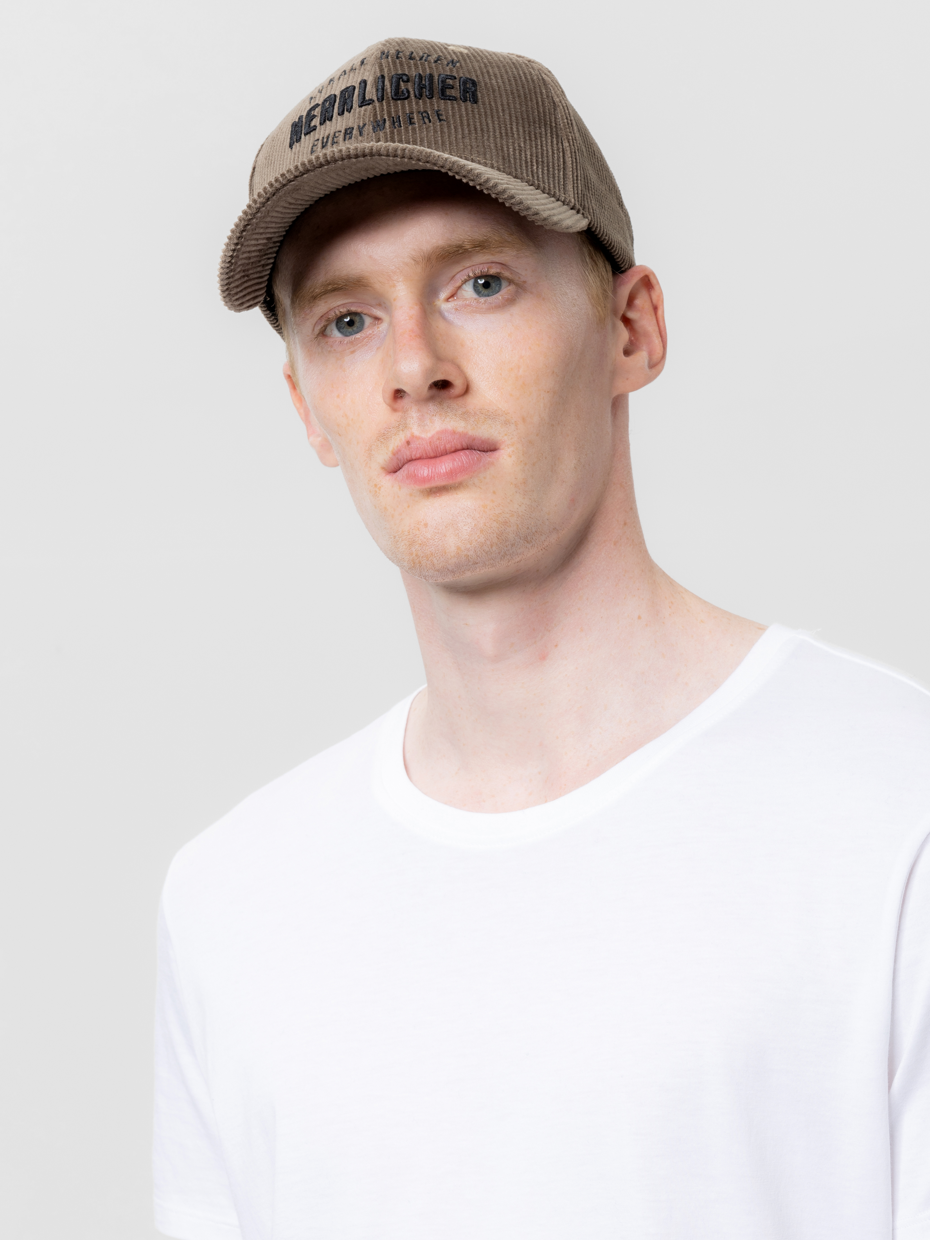 Ein junger Mann mit heller Haut trägt eine hellbraune Baseballkappe mit schwarzer Aufschrift und ein schlichtes weißes T-Shirt vor einem hellgrauen Hintergrund.