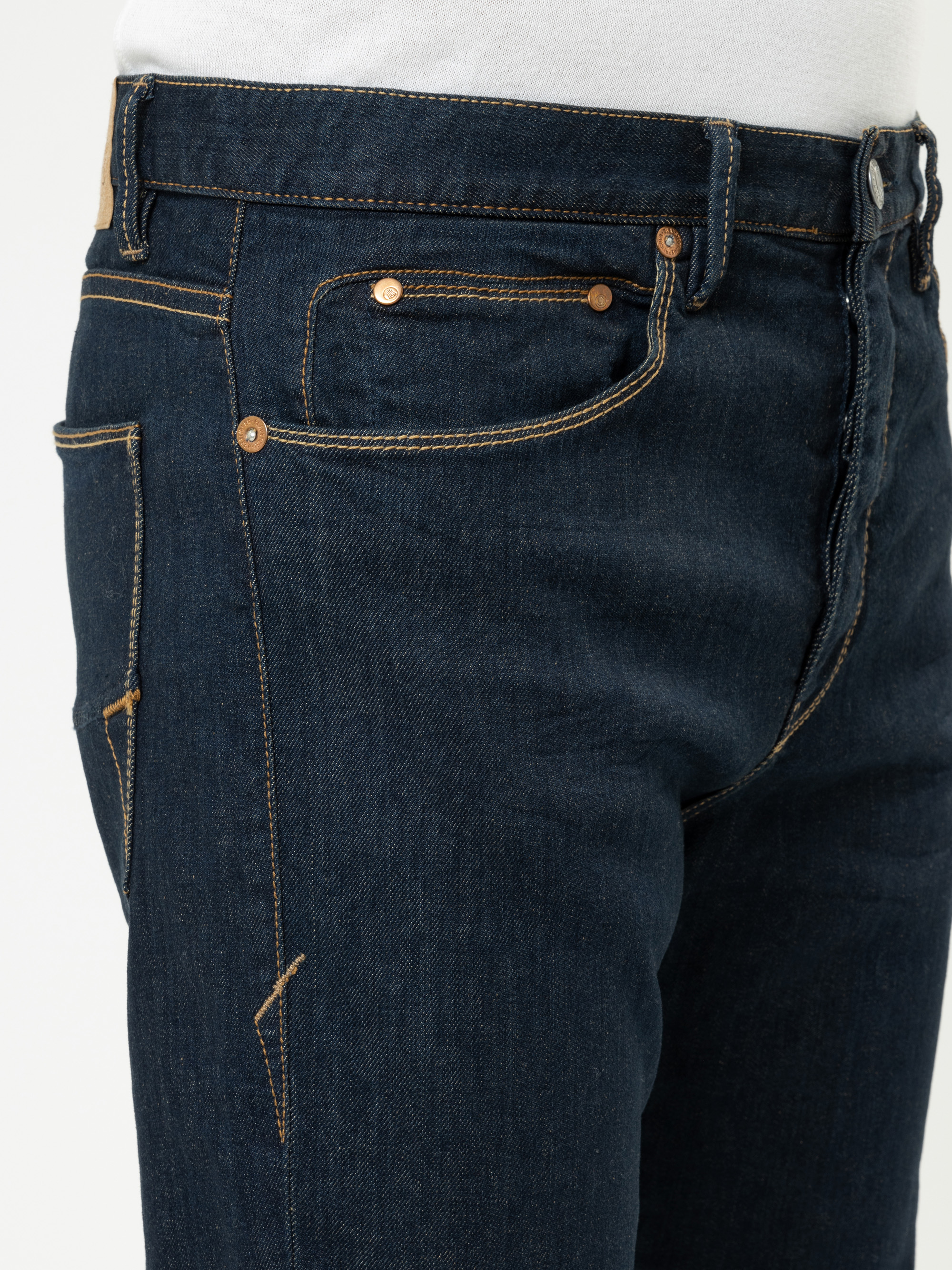 Nahaufnahme einer Person, die eine dunkelblaue Jeans mit kupferfarbenen Nieten und gelben Nähten trägt, gepaart mit einem weißen Hemd, das in der Taille gesteckt wird.
