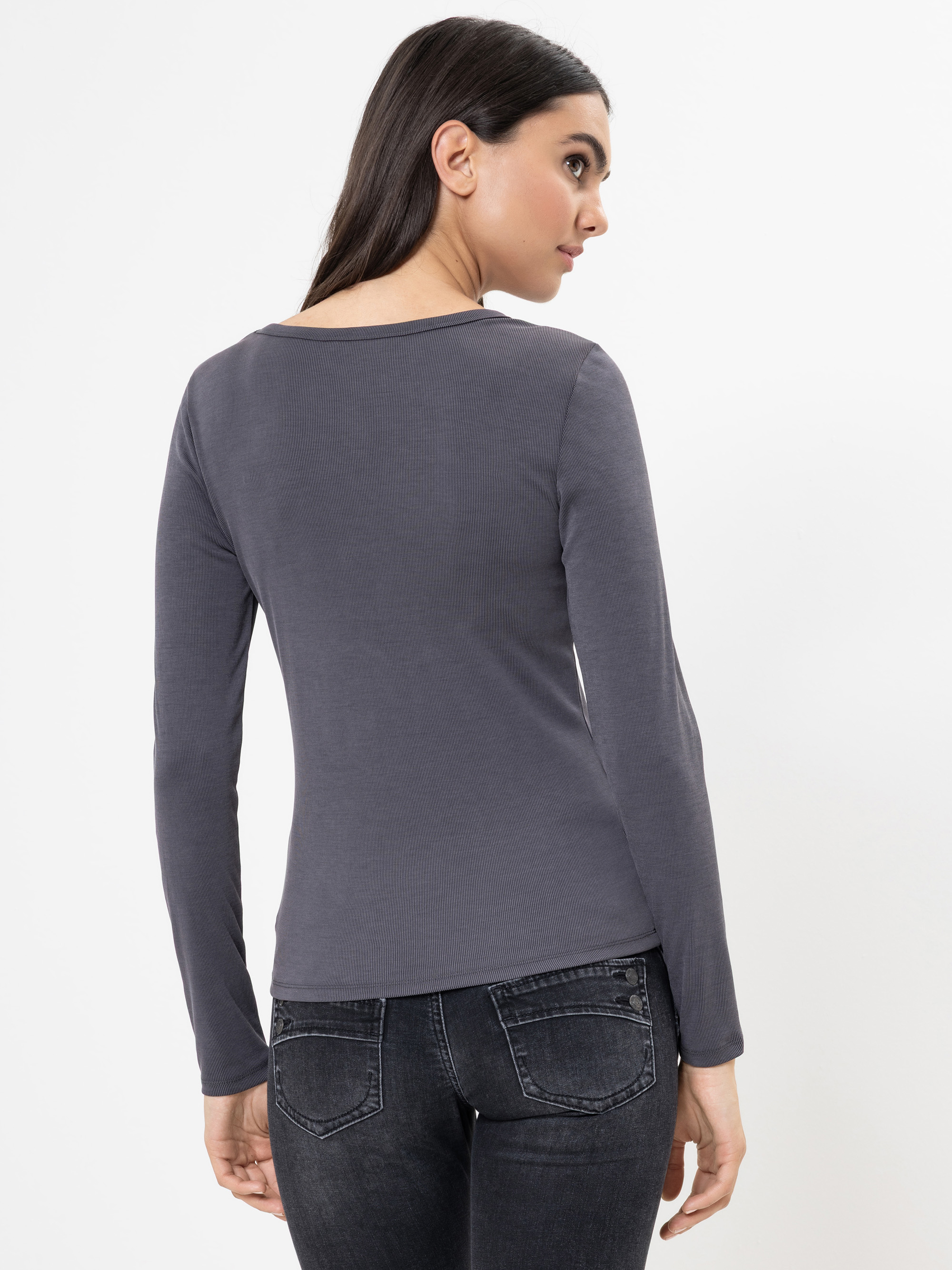 Eine Frau mit langen dunklen Haaren, bekleidet mit einem taillierten, langärmeligen, anthrazitfarbenen Oberteil und dunkelgrauen Jeans, steht mit dem Blick zur Seite und blickt über ihre Schulter auf einen schlichten weißen Hintergrund.