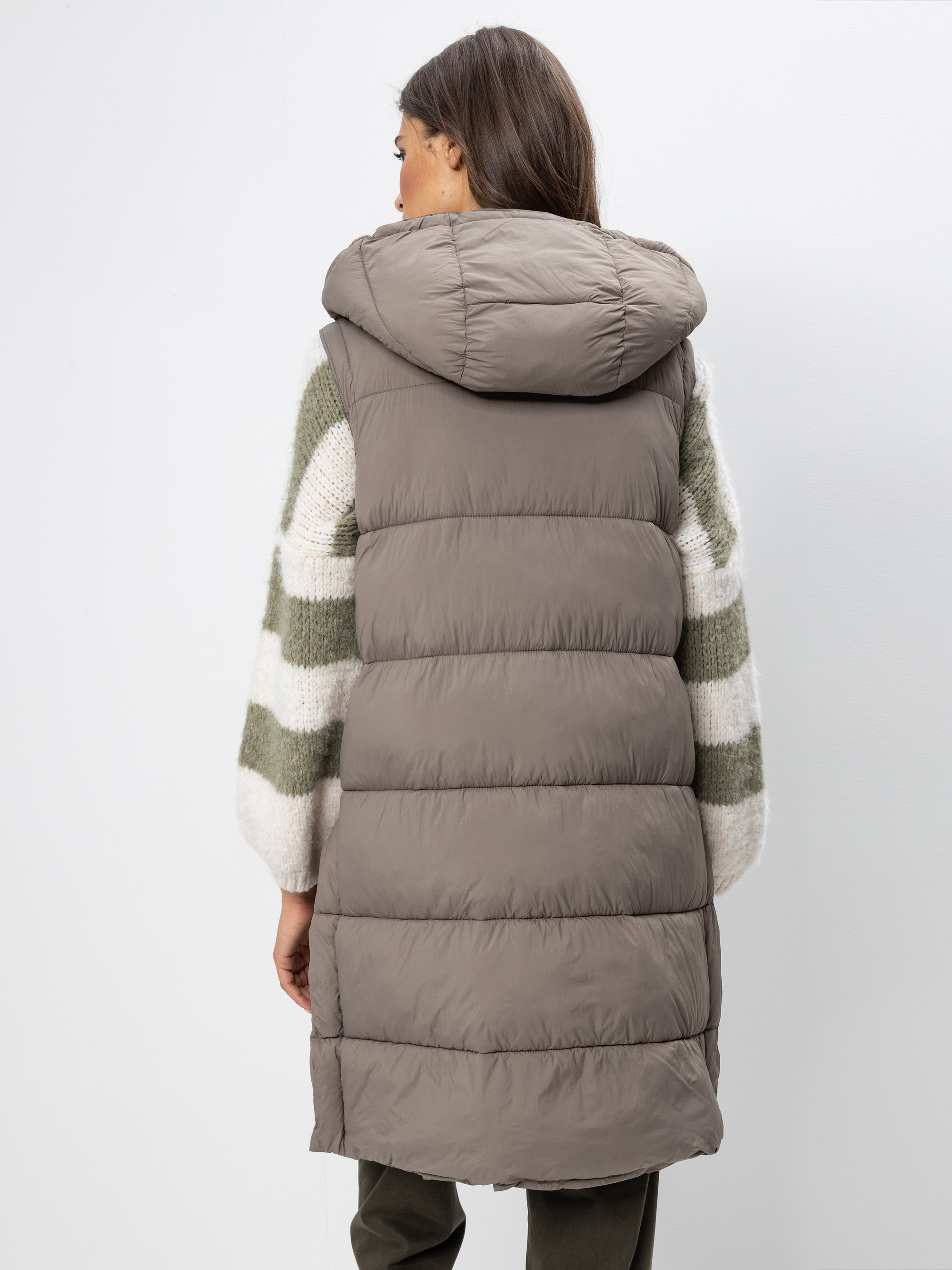 Eine Frau mit langen braunen Haaren ist von hinten zu sehen, die eine bauschige, taupefarbene Kapuzenweste über einem cremefarbenen Pullover mit grünen Streifen und eine dunkle Hose trägt und vor einem schlichten weißen Hintergrund steht.