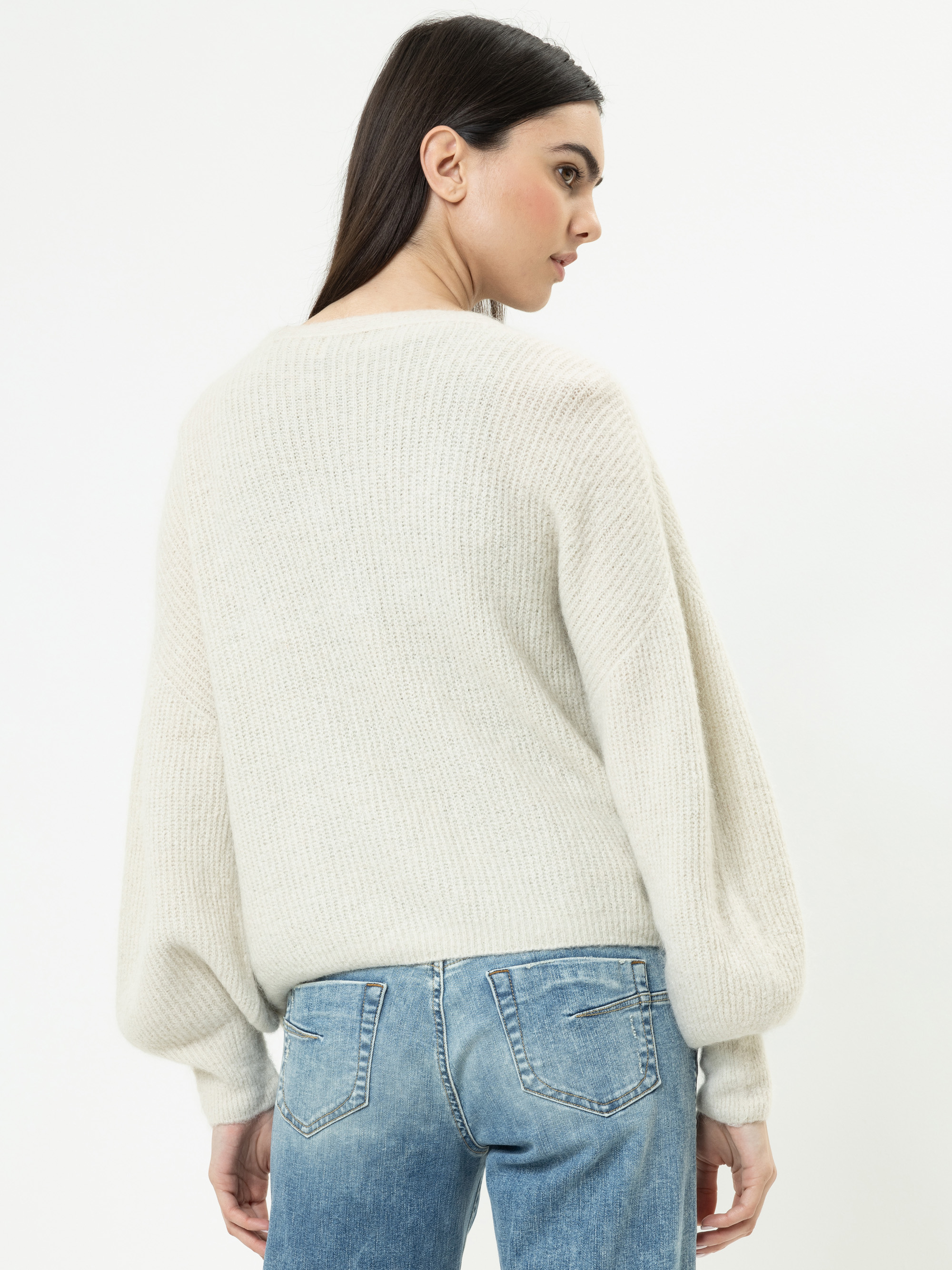 Eine Frau mit langen dunklen Haaren trägt einen cremefarbenen Strickpullover und blaue Jeans. Sie steht und schaut leicht von der Kamera weg, so dass die Rückseite ihres Outfits vor einem schlichten weißen Hintergrund zu sehen ist.