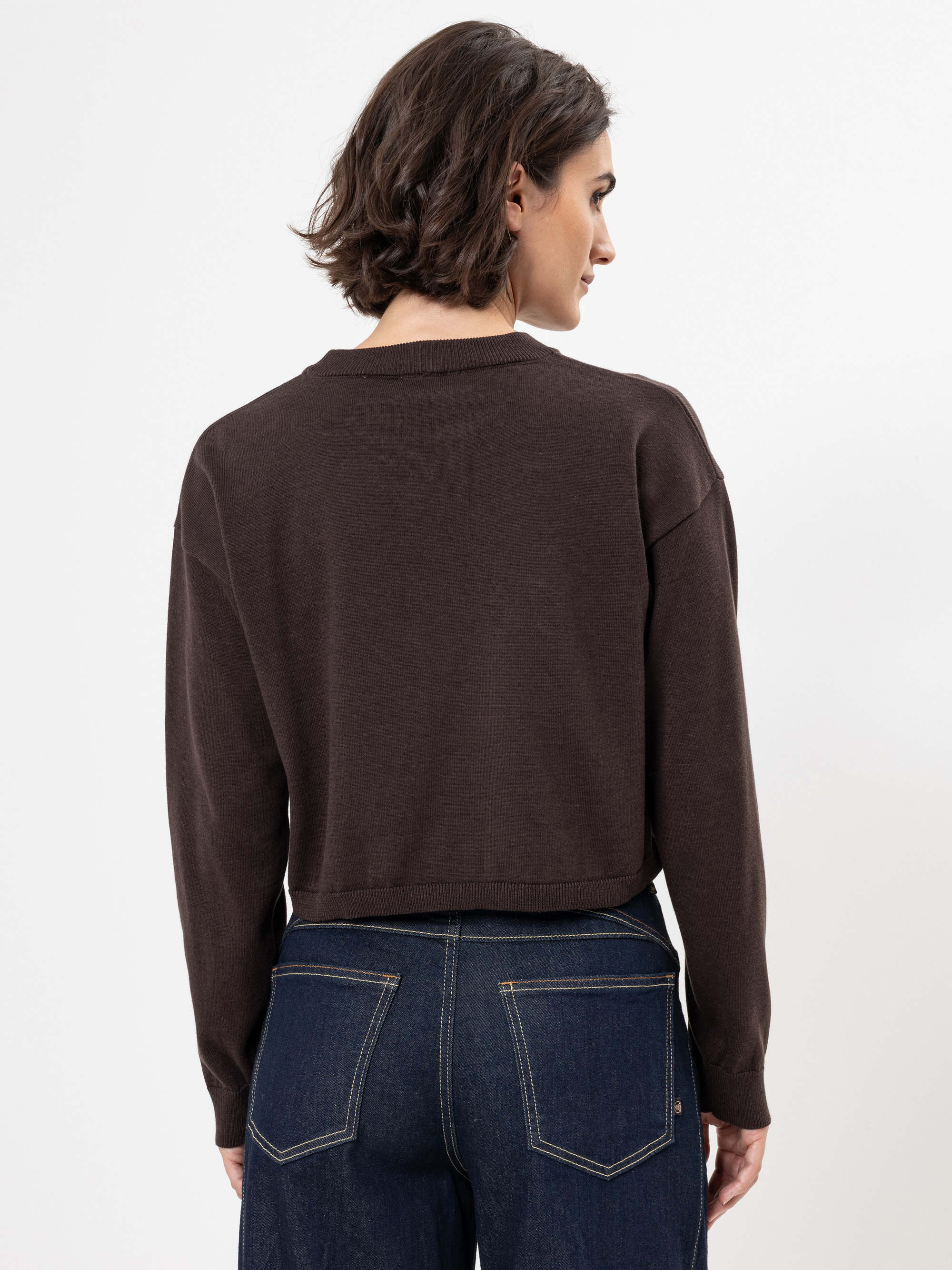 Eine Frau mit kurzen braunen Haaren, bekleidet mit einem dunkelbraunen, langärmeligen Pullover und hoch sitzenden Bluejeans, steht vor einem schlichten weißen Hintergrund und blickt von der Kamera weg.