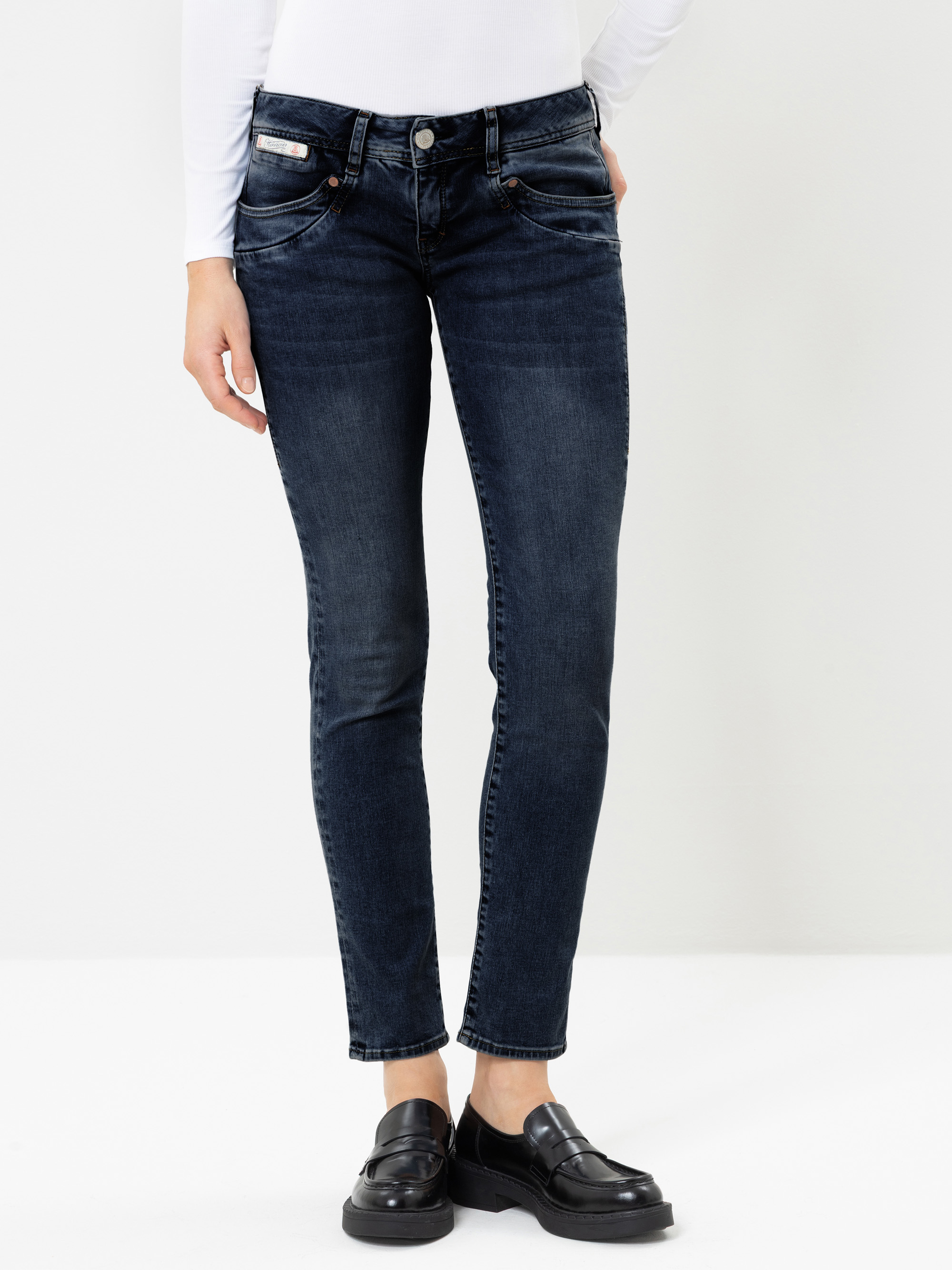 Eine Person, die eine dunkelblaue, schmal geschnittene Jeans, ein weißes Langarm-Oberteil und schwarze Slipper trägt und vor einem schlichten weißen Hintergrund steht. Der Kopf ist auf dem Bild nicht zu sehen.