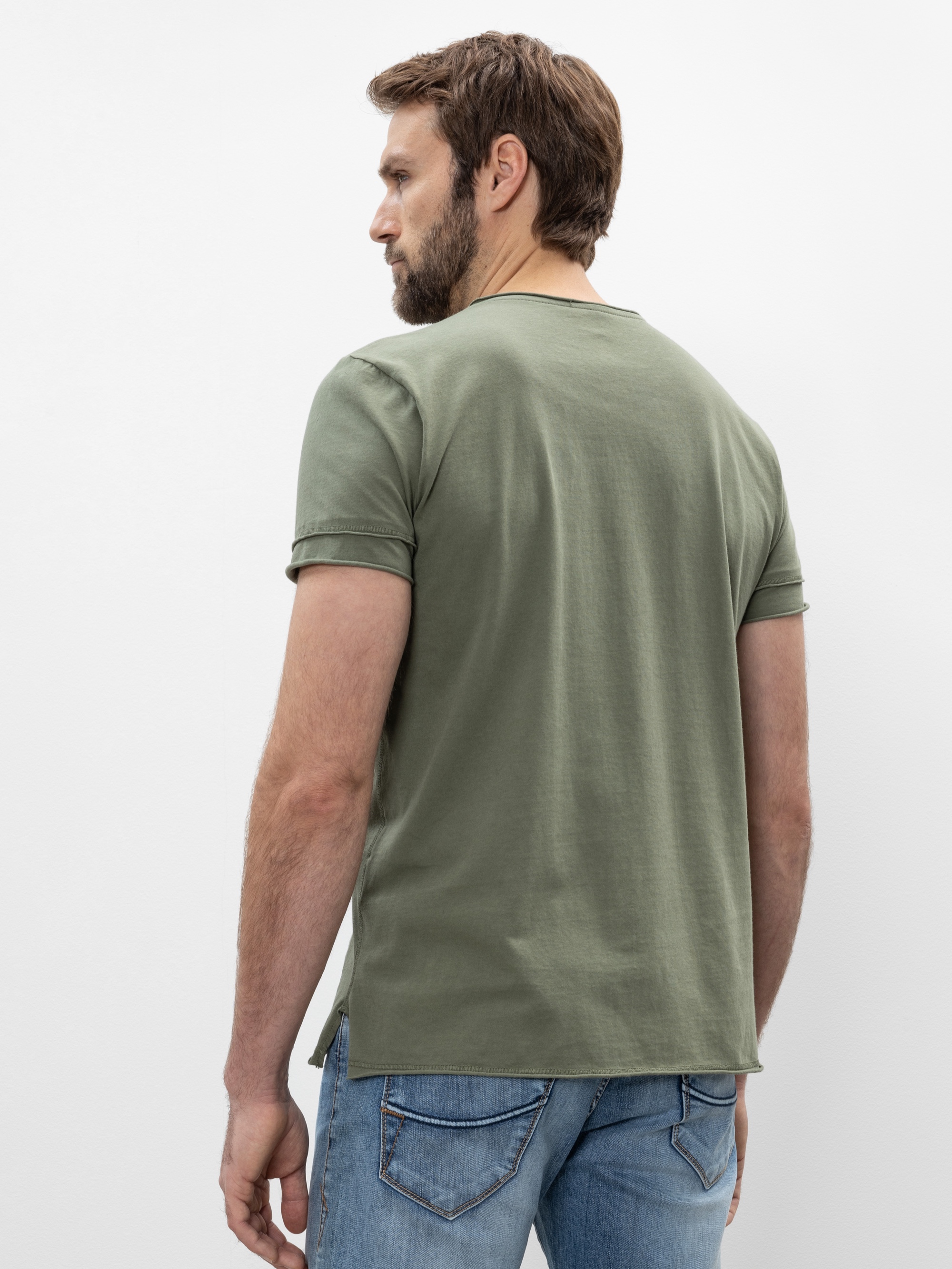 Ein Mann mit hellbraunem Haar und Bart steht mit dem Rücken zur Kamera, trägt ein salbeigrünes T-Shirt und hellblaue Jeans vor einem schlichten weißen Hintergrund.