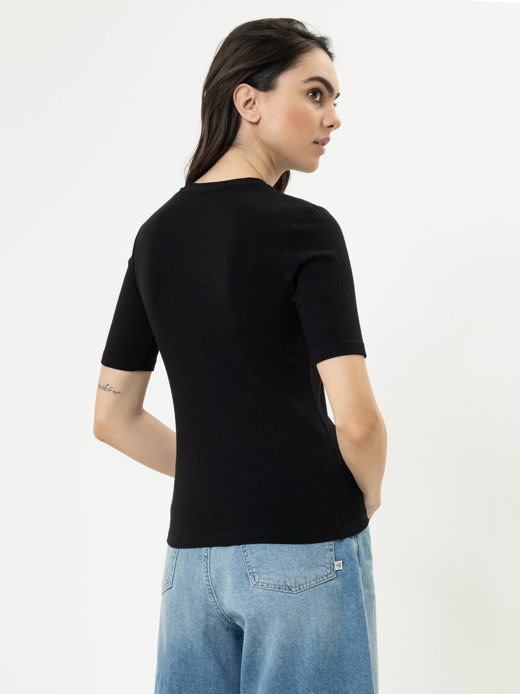 Eine Frau mit langen braunen Haaren, die ein schwarzes geripptes kurzärmeliges Oberteil und hellblaue Jeans trägt, steht leicht von der Kamera abgewandt vor einem schlichten weißen Hintergrund. Auf ihrem linken Unterarm ist eine kleine Tätowierung zu