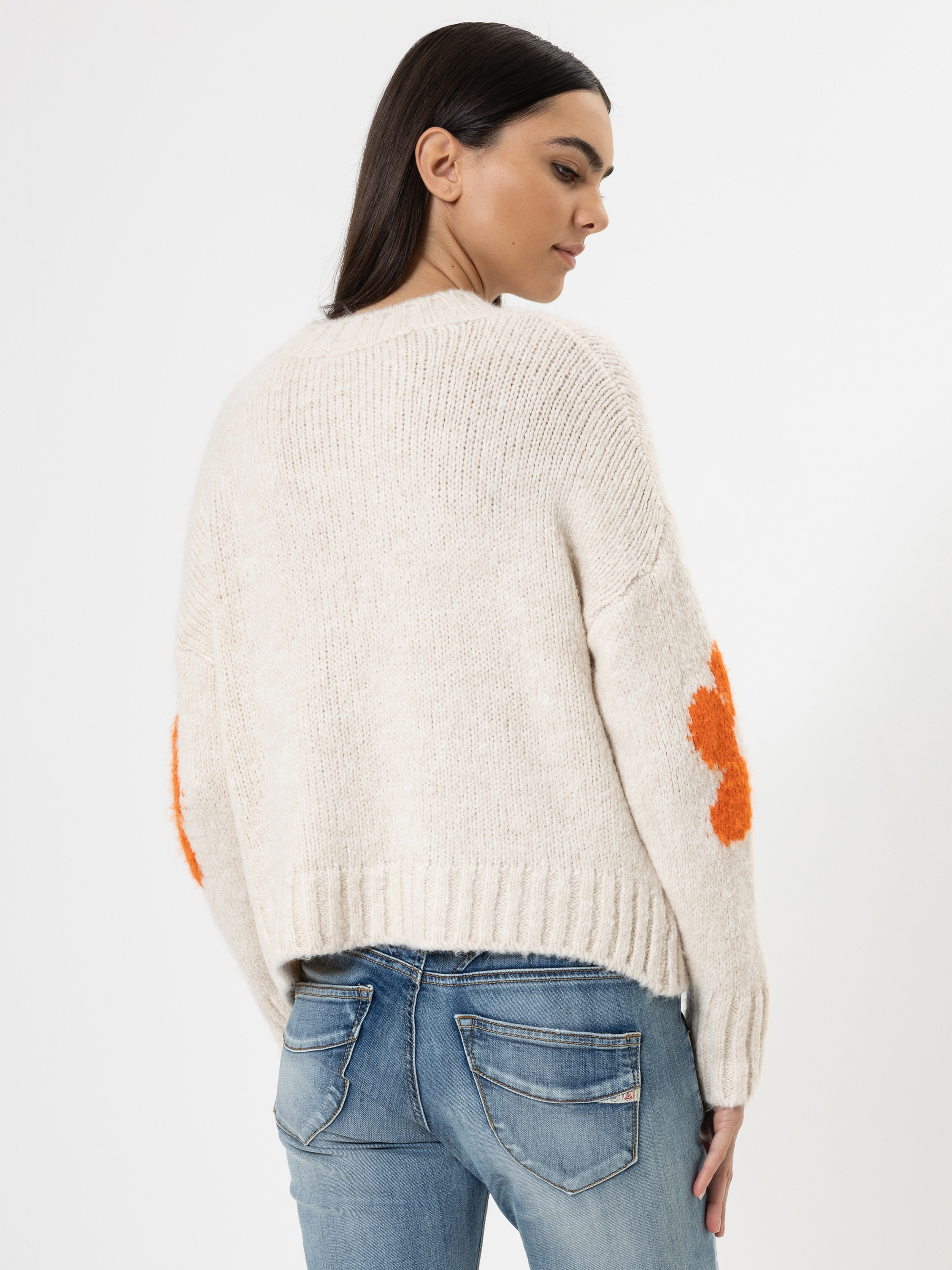 Eine Frau mit glattem dunklem Haar trägt einen cremefarbenen Strickpullover mit orangefarbenen Blumenaufnähern an den Ärmeln und blaue Jeans. Sie steht mit dem Rücken zur Kamera vor einem weißen Hintergrund.