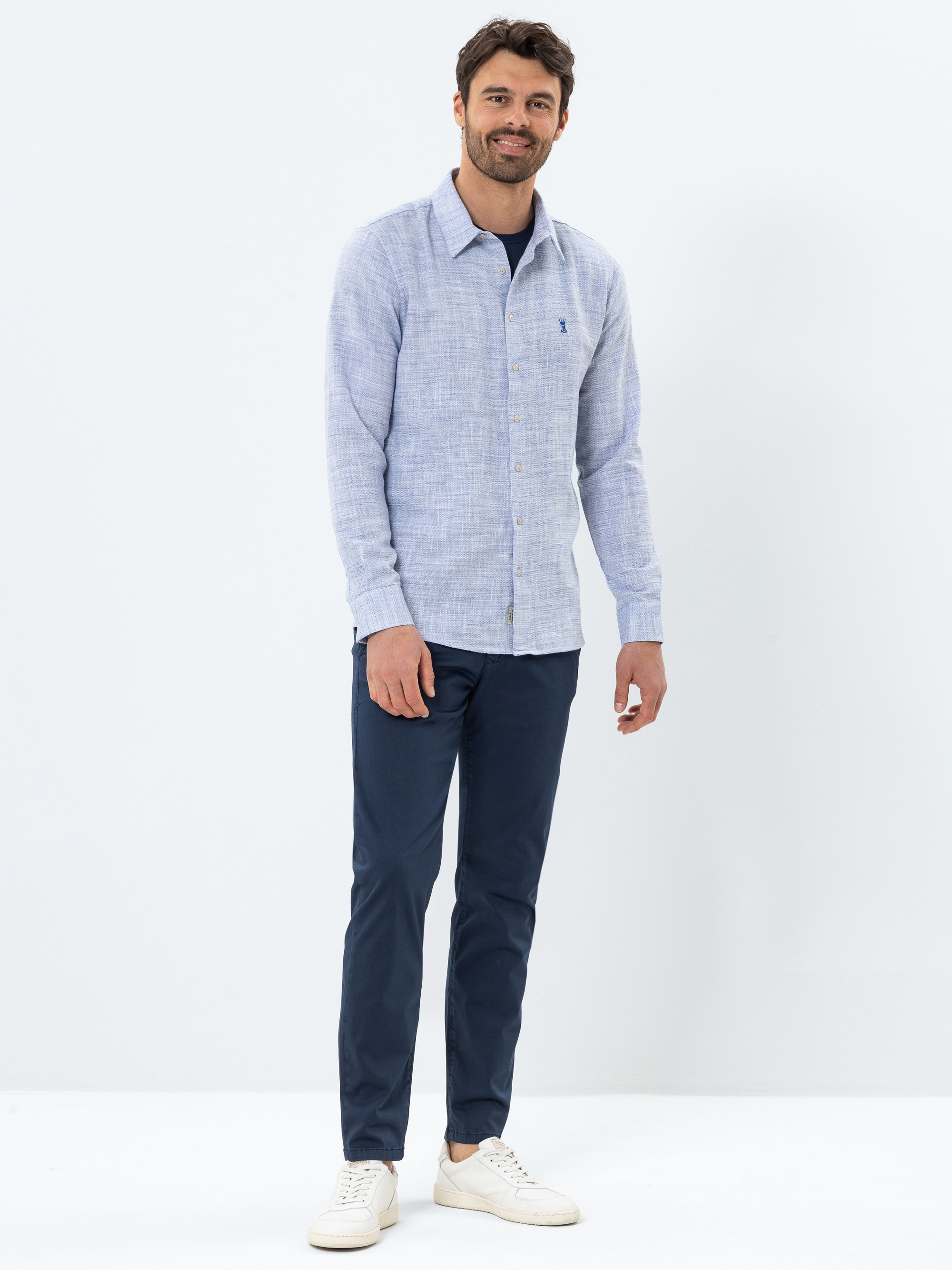 Ein Mann, der ein hellblaues Button-up-Hemd über einem marineblauen T-Shirt, eine marineblaue Hose und weiße Turnschuhe trägt, steht lächelnd vor einem schlichten weißen Hintergrund.