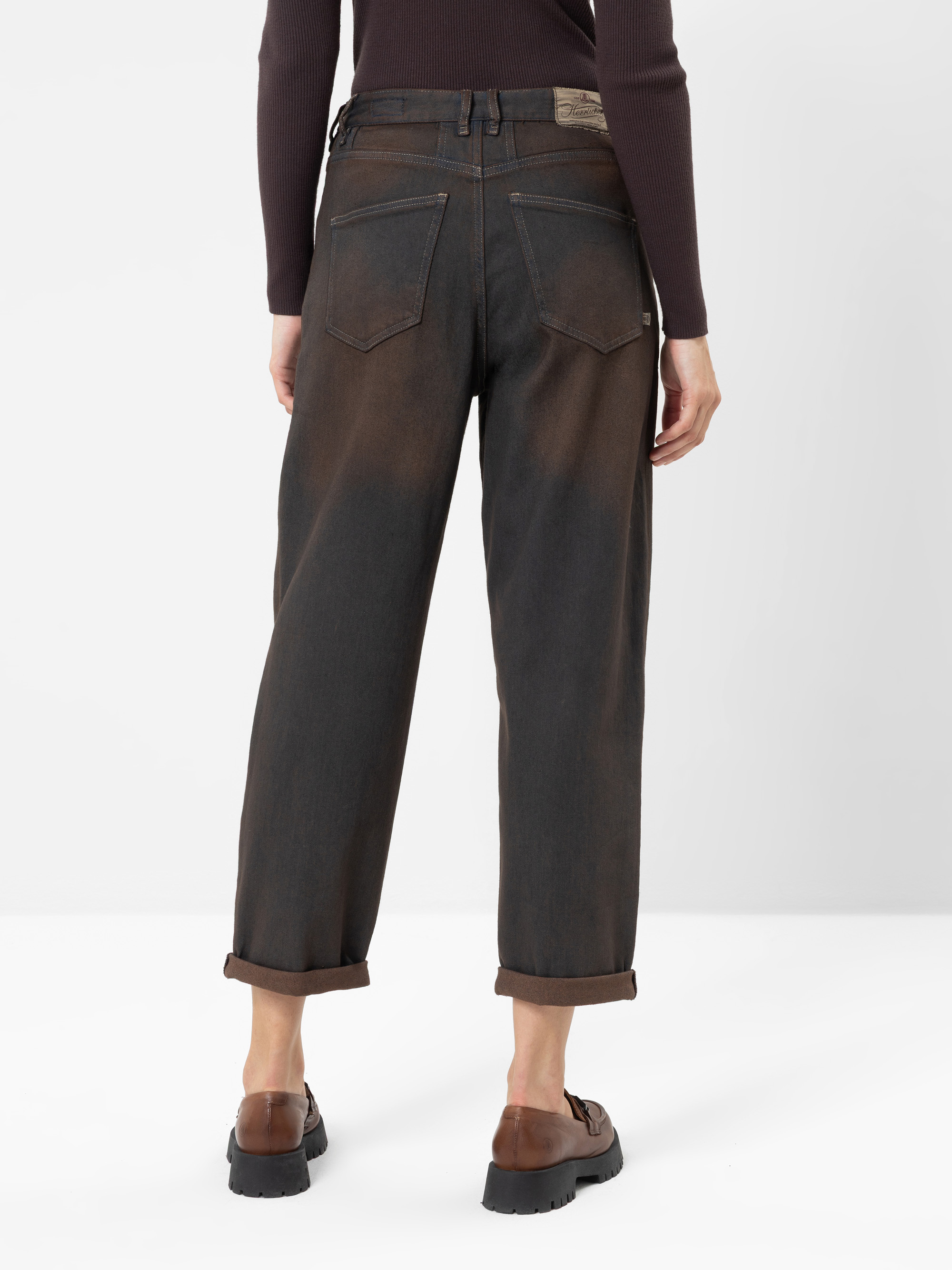 Eine Person, die eine dunkelbraune Jeans mit Umschlag, braune Schuhe und ein dunkles Langarmshirt trägt, von hinten vor einem einfarbigen weißen Hintergrund gesehen.