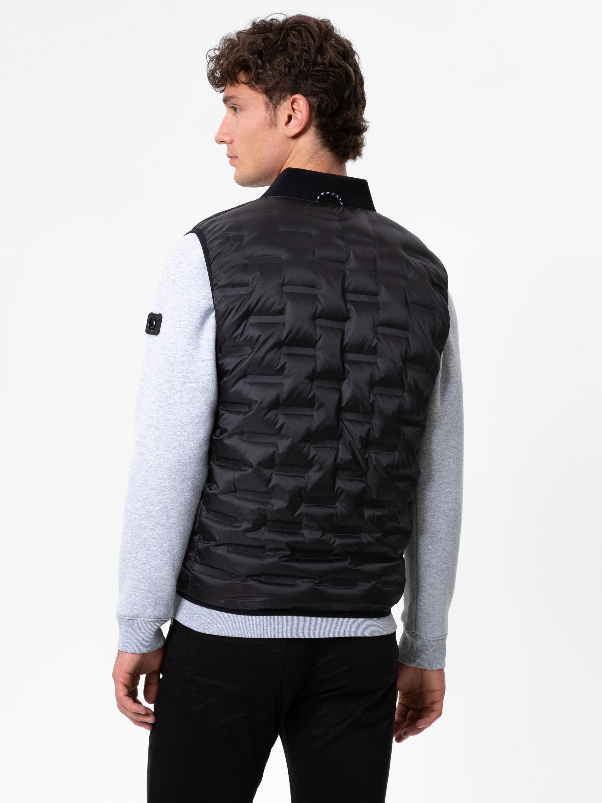 Ein Mann mit kurzen lockigen Haaren wird von hinten gezeigt. Er trägt eine schwarze Steppweste über einem hellgrauen langärmeligen Hemd und eine schwarze Hose und steht vor einem schlichten weißen Hintergrund.