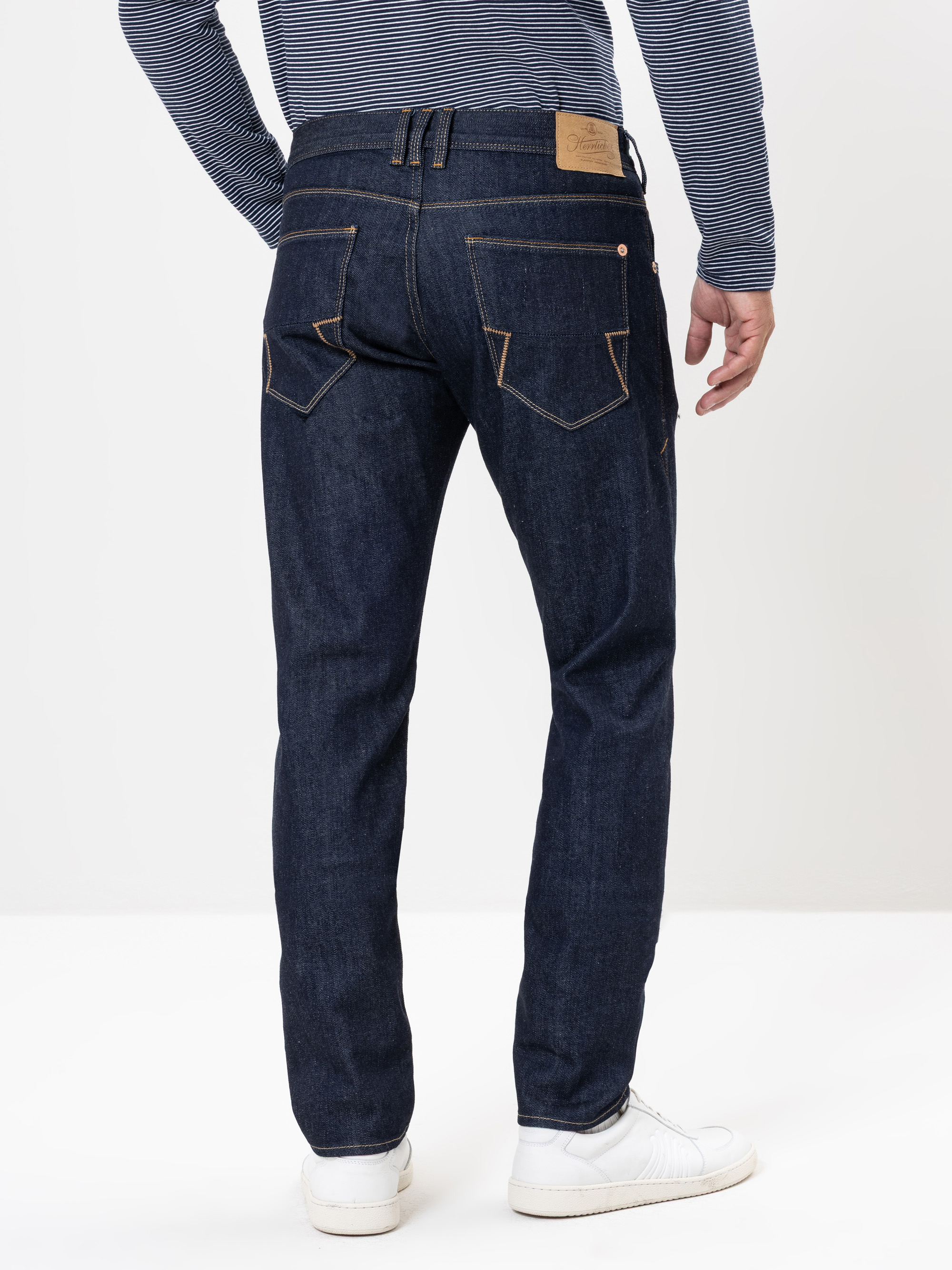 Rückenansicht einer Person, die dunkelblaue Jeans, ein blau-weiß gestreiftes langärmeliges Hemd und weiße Turnschuhe trägt und vor einem schlichten weißen Hintergrund steht.