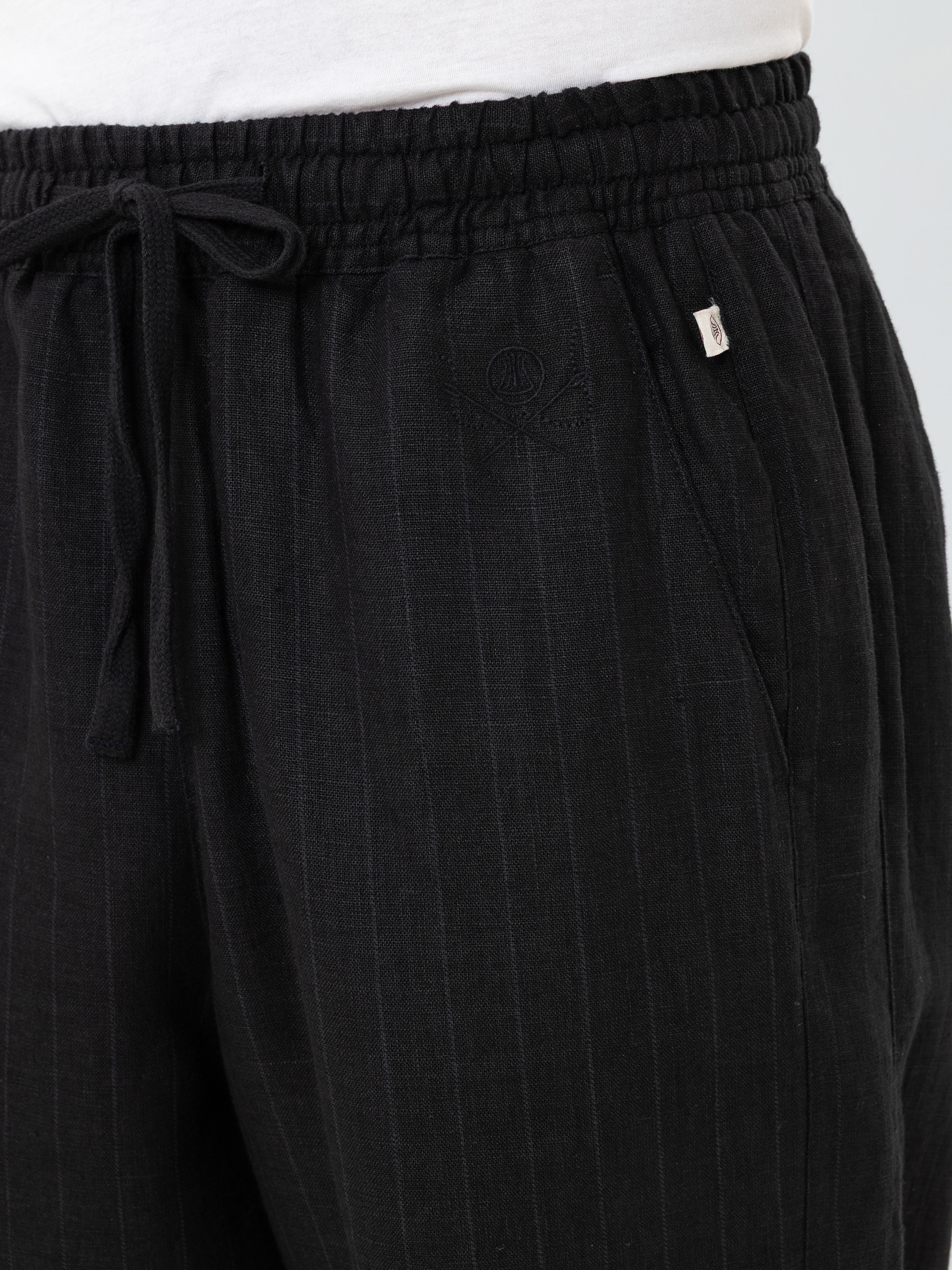Nahaufnahme einer Person, die eine schwarze Hose mit Kordelzug und dezenten Nadelstreifen, Vordertaschen und einem kleinen gestickten Logo in der Nähe der linken Tasche trägt, kombiniert mit einem weißen Hemd.