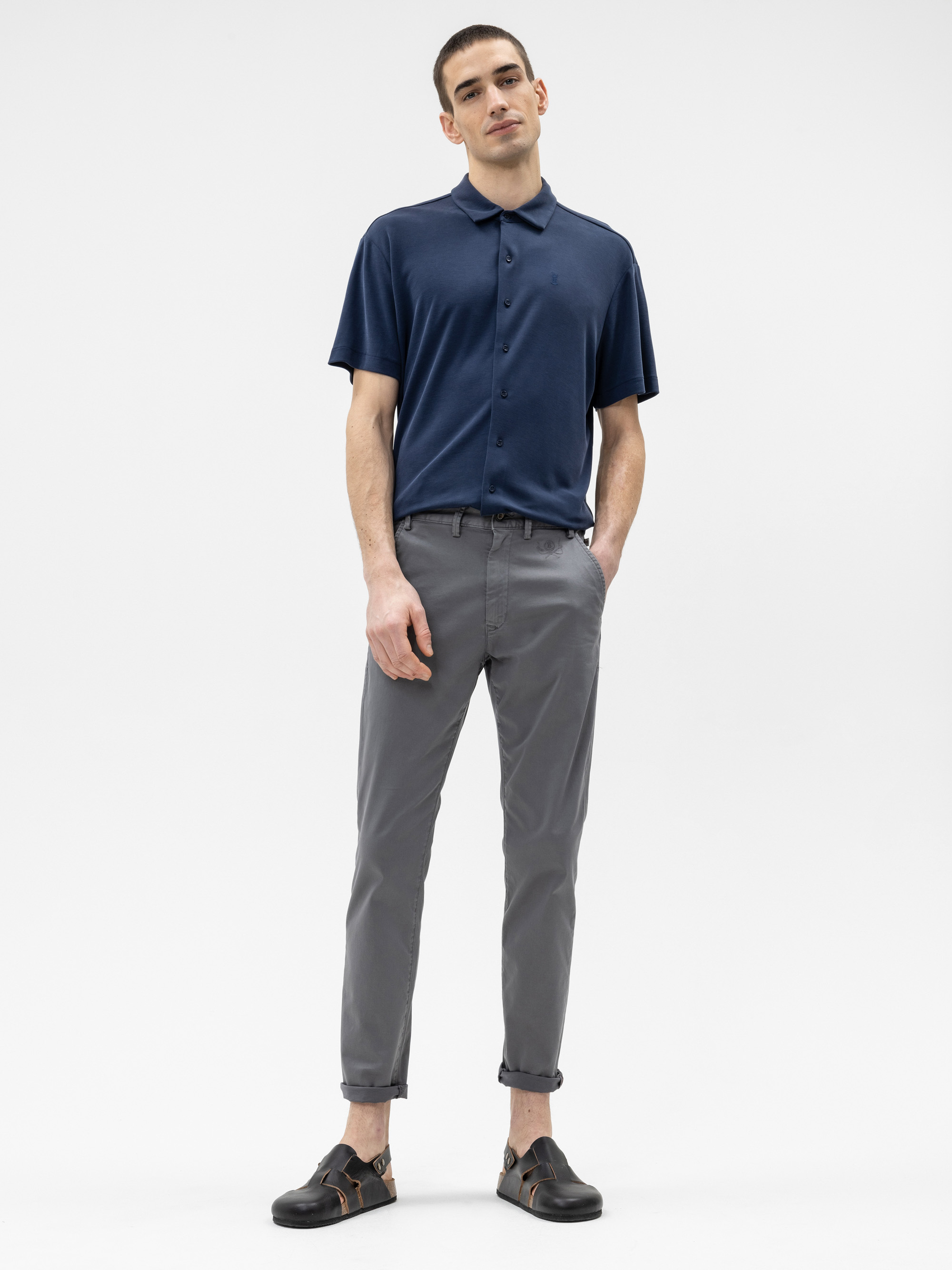 Ein Mann steht vor einem schlichten weißen Hintergrund, trägt ein marineblaues, kurzärmeliges Polohemd, eine graue Hose mit Bündchen und schwarze Sandalen. Seine Hände stecken in den Hosentaschen, und er blickt selbstbewusst in die Kamera.