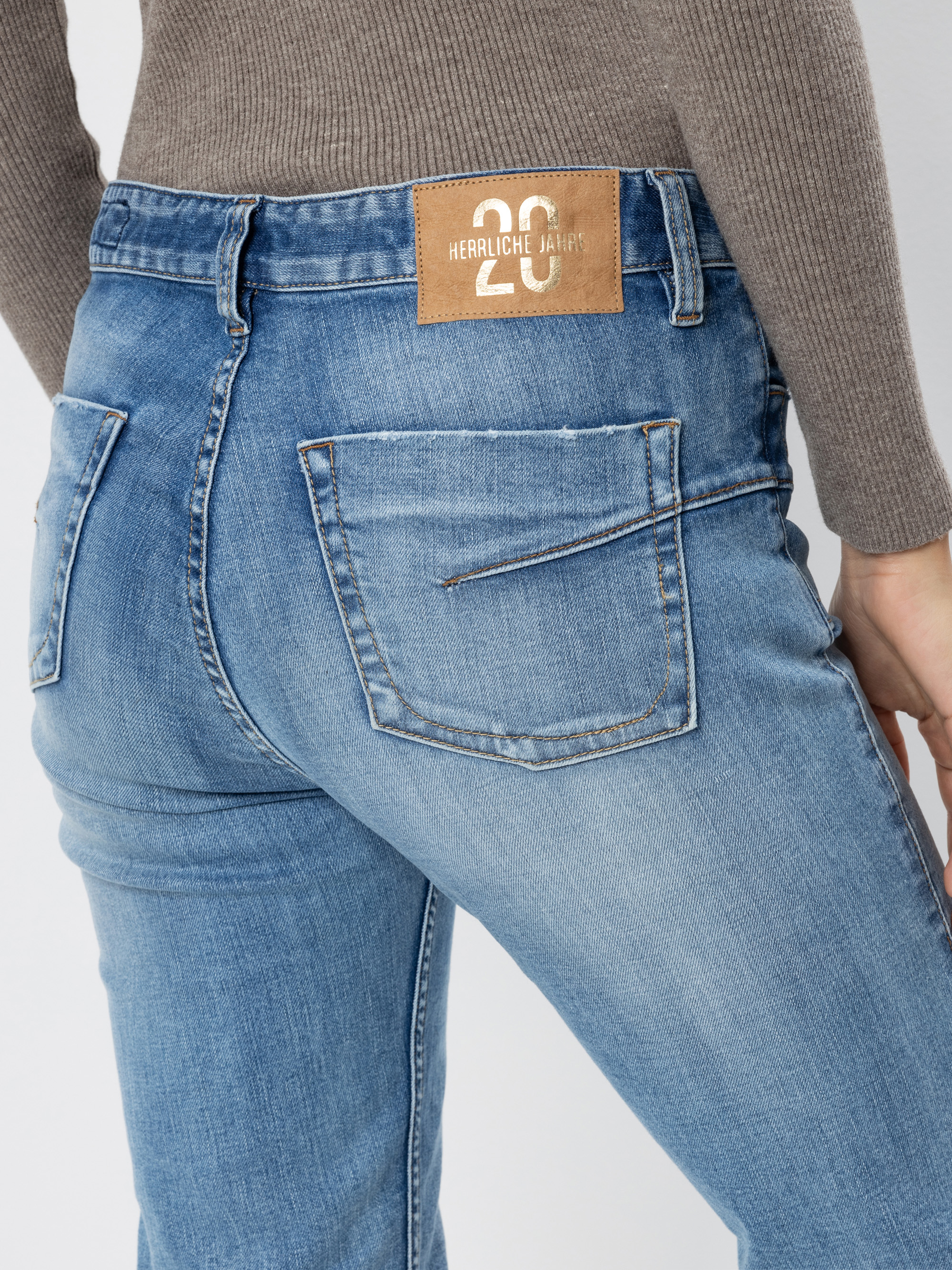 Eine Person trägt eine hellblaue Jeans und ein geripptes graues Oberteil, das von der Taille abwärts und von hinten gezeigt wird. Die Jeans hat einen braunen Lederaufnäher mit weißem Text am Bund und sichtbare Gesäßtaschen.