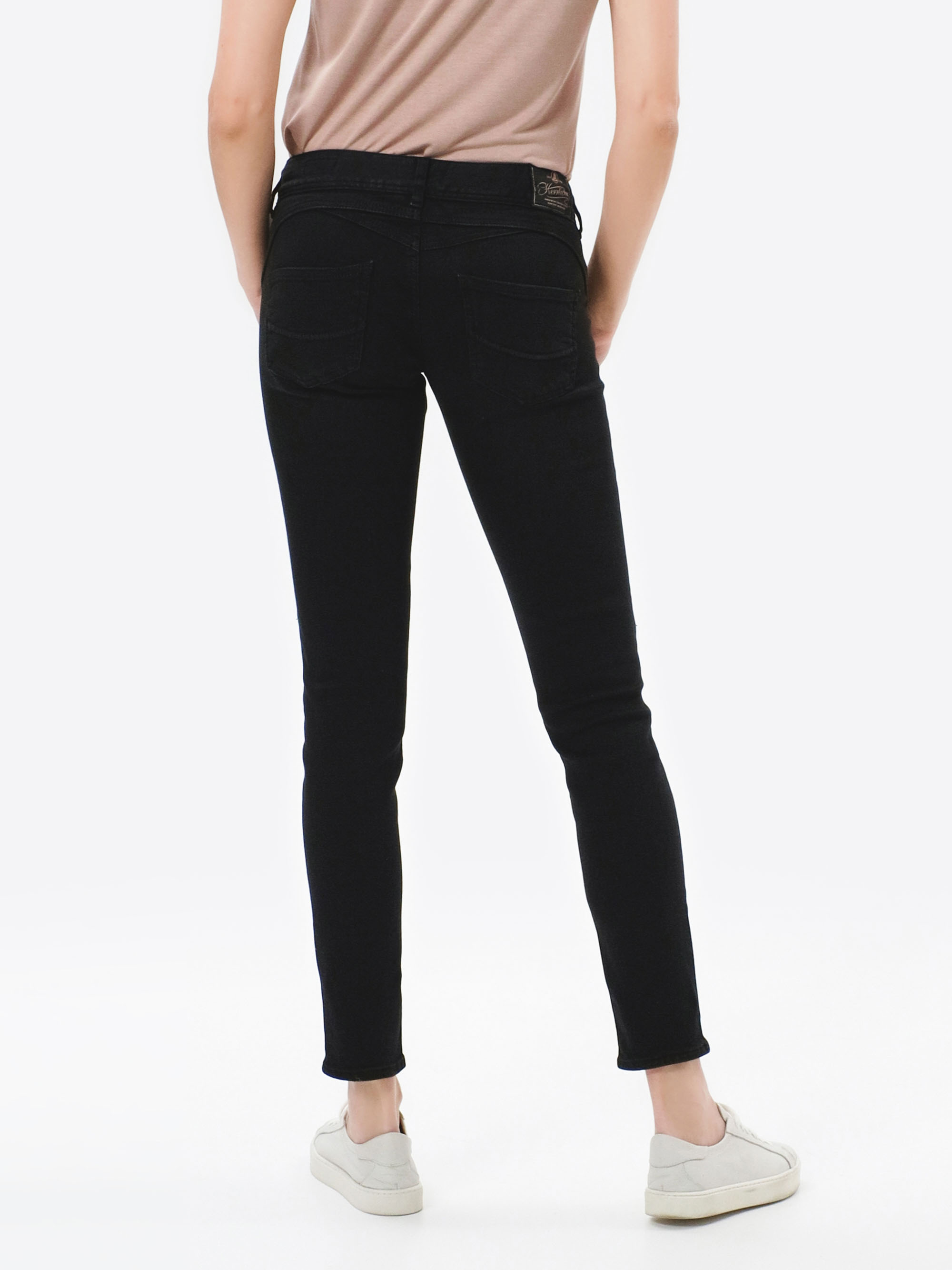 Eine Person, die ein beiges Oberteil, schwarze Röhrenjeans und weiße Turnschuhe trägt, steht mit dem Rücken zur Kamera vor einem schlichten weißen Hintergrund.