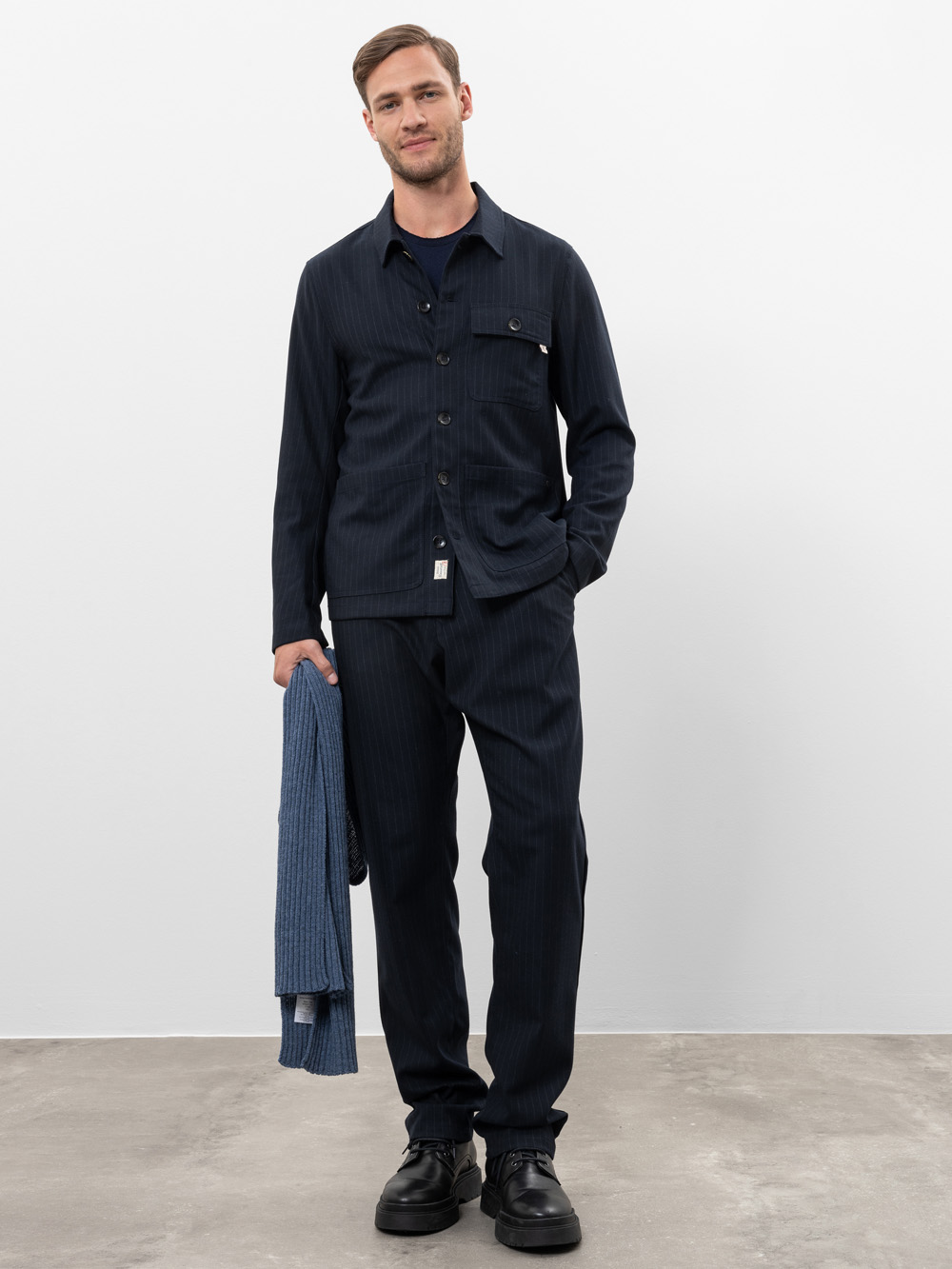 Ein Mann steht auf einem Betonboden vor einer schlichten weißen Wand, trägt eine dunkle Nadelstreifenjacke und eine dazu passende Hose, hält einen gefalteten blauen Pullover in der linken Hand und lächelt leicht in die Kamera.