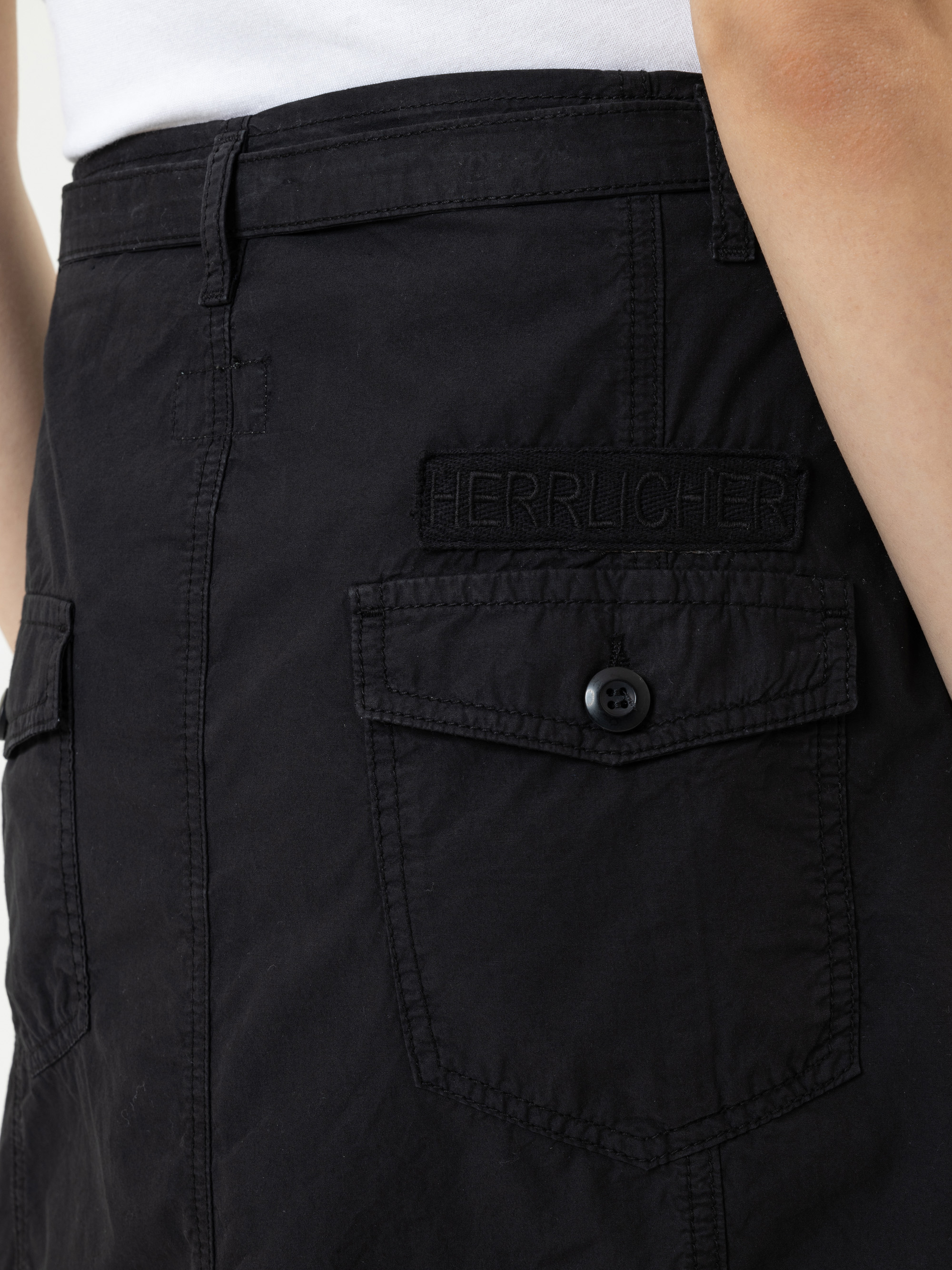Nahaufnahme einer Person, die eine schwarze Hose mit einer Pattentasche mit einem Knopf und einem aufgestickten HERRLICHER-Etikett über der Tasche trägt. Die Person trägt außerdem ein weißes Oberteil.